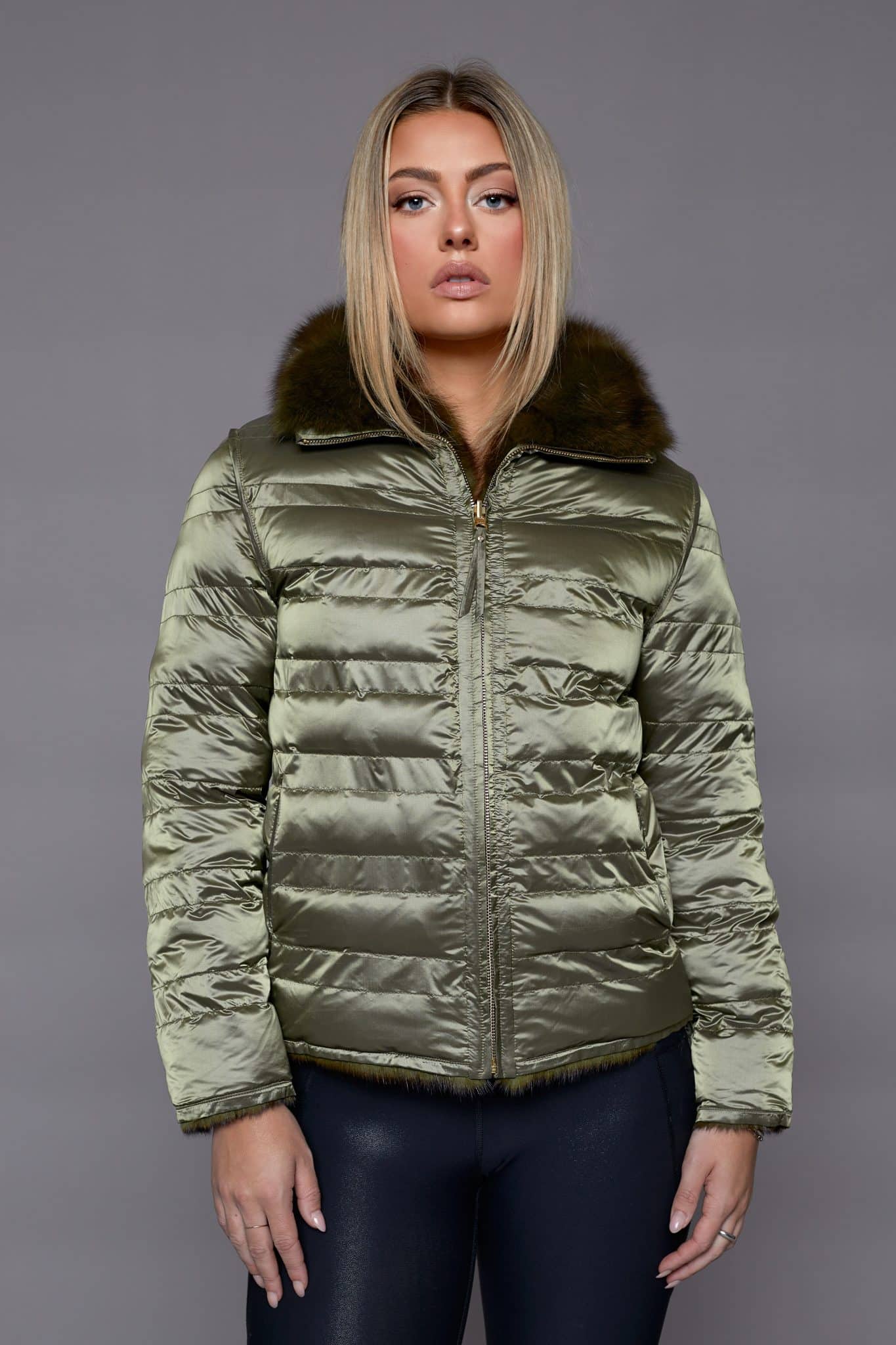 Frontal view of a woman wearing a reversible fur coat with the shell side out standing in front of a grey background.