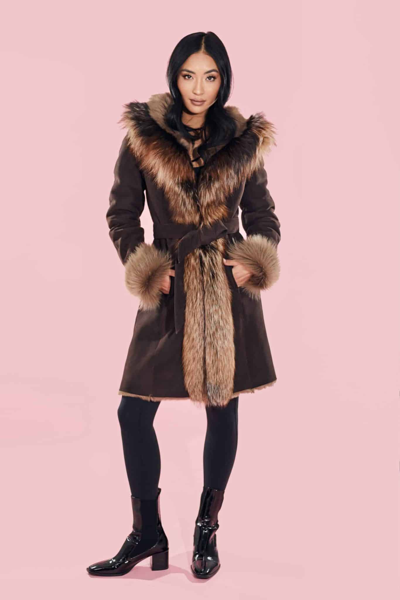 Frontal view of a woman wearing a fur coat with the hood down standing in front of a pink background.