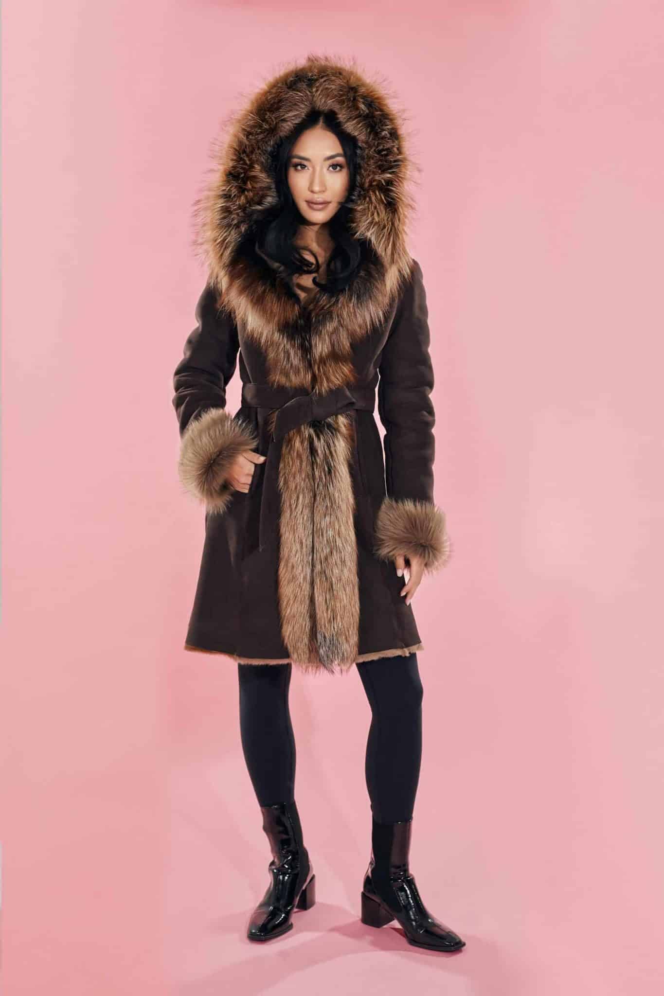 Frontal view of a woman wearing a fur coat with the hood up standing in front of a pink background.