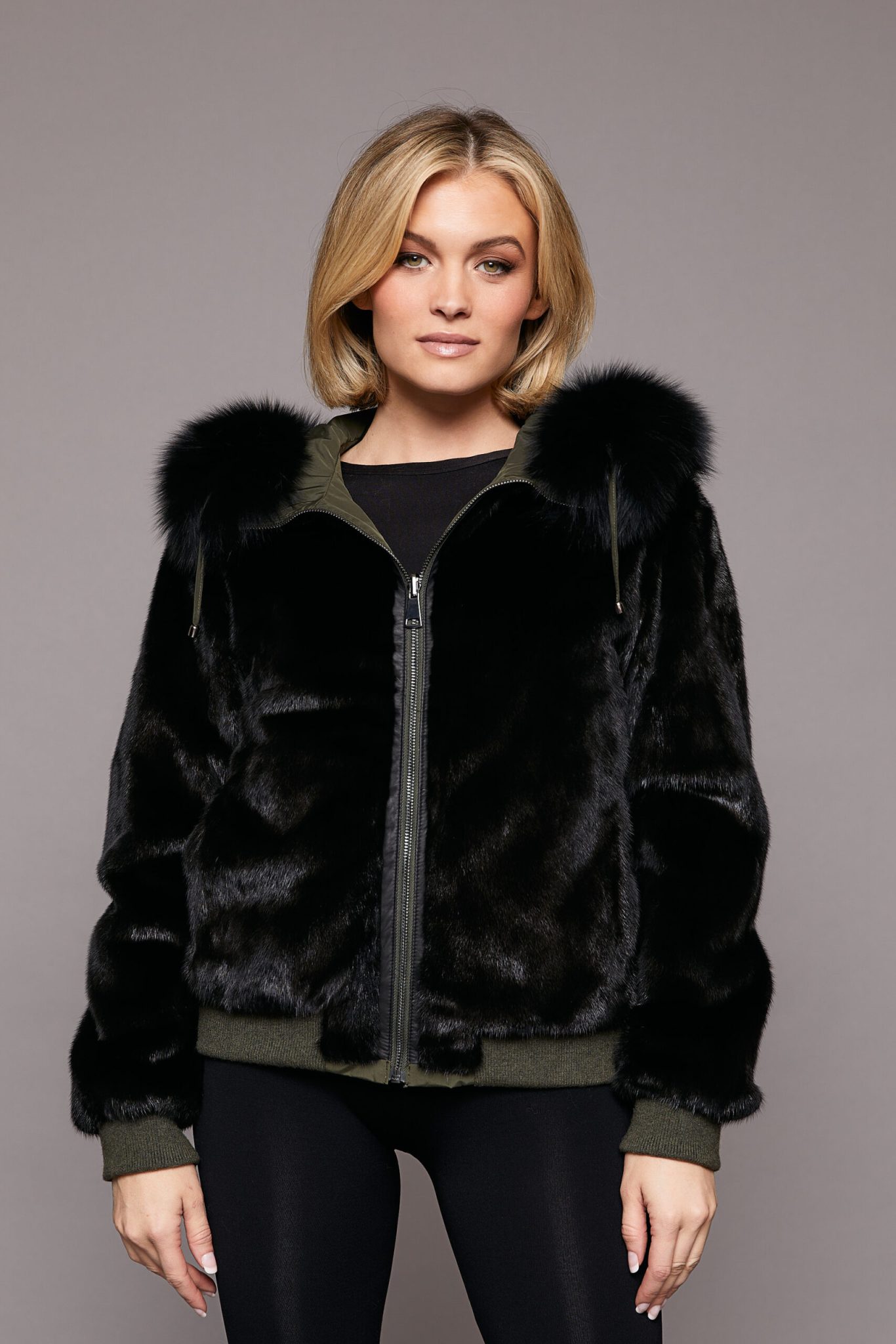 Frontal view of a woman wearing a black fur coat with the hood down and fur side out standing in front of a grey background. The woman's hands are positioned at her sides.
