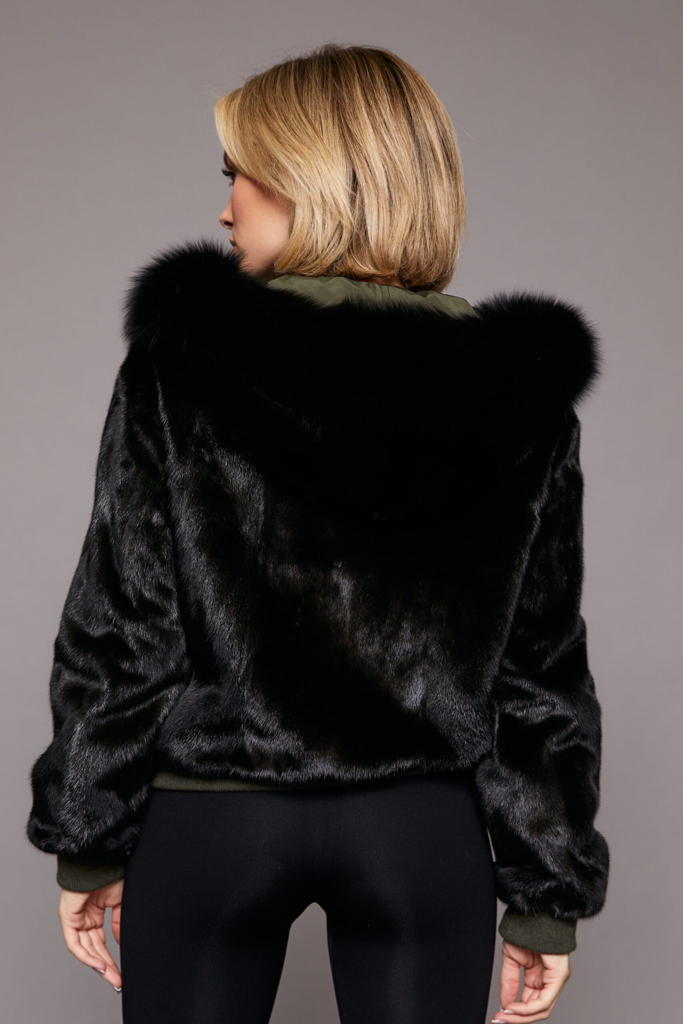 Rear view of a woman wearing a black fur coat with the hood down and fur side out facing a grey background.