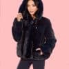 Frontal view of a woman wearing a fur coat with the hood up standing in front of a pink background.
