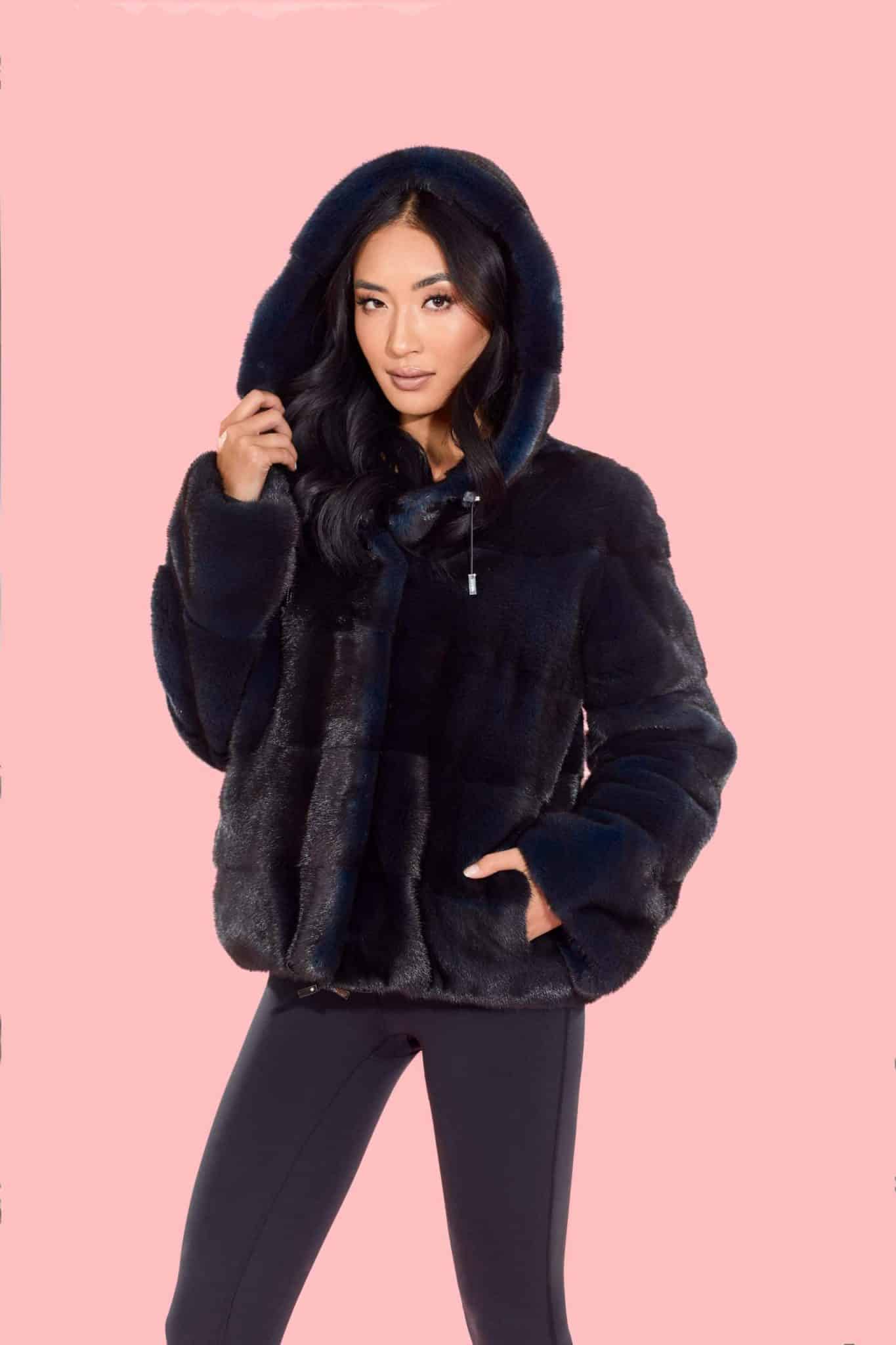 Frontal view of a woman wearing a fur coat with the hood up standing in front of a pink background.