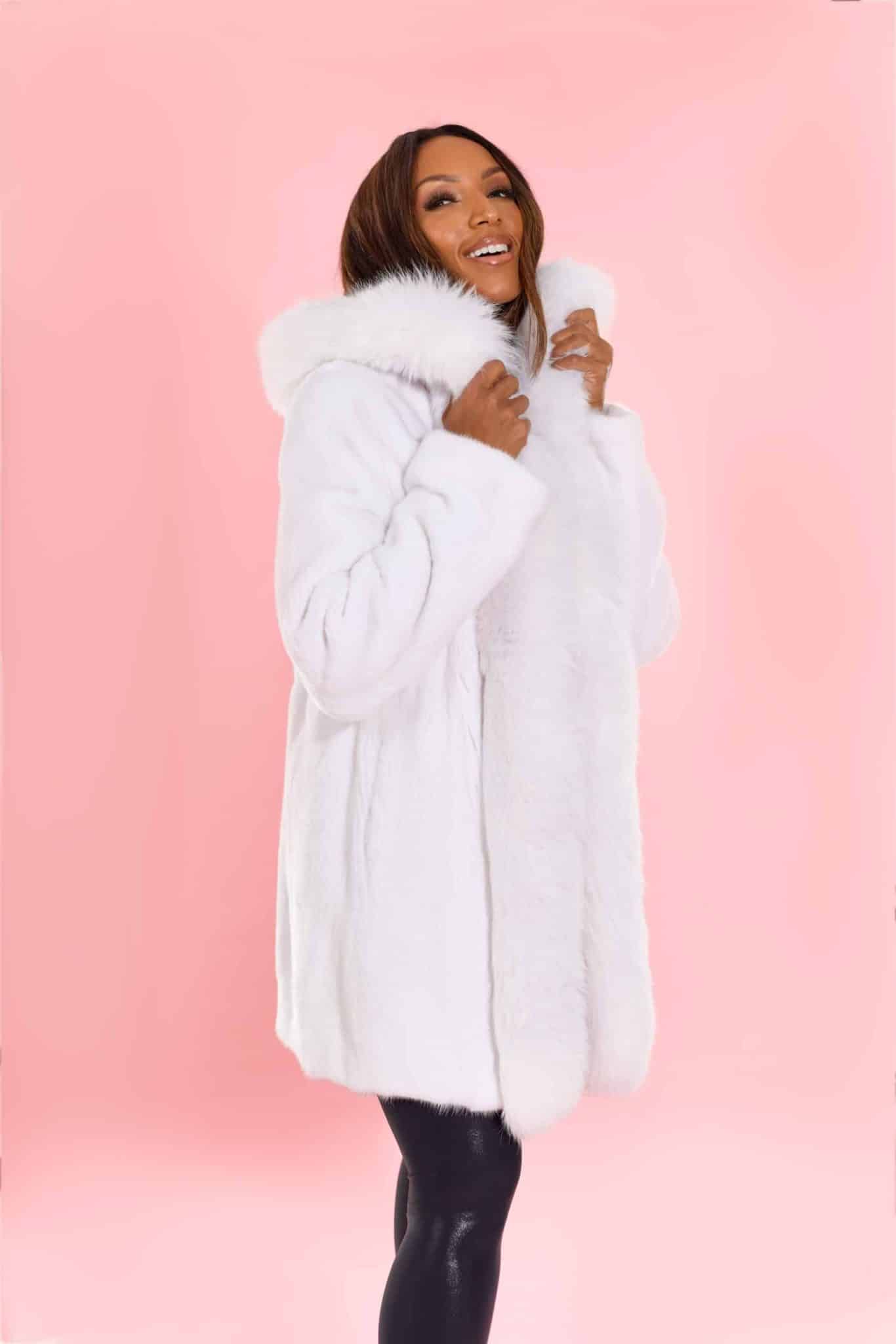 Profile view of a woman wearing a white mink coat with the hood down standing in front of a pink background. The woman's hands are clutching the bottom of the coat's hood.
