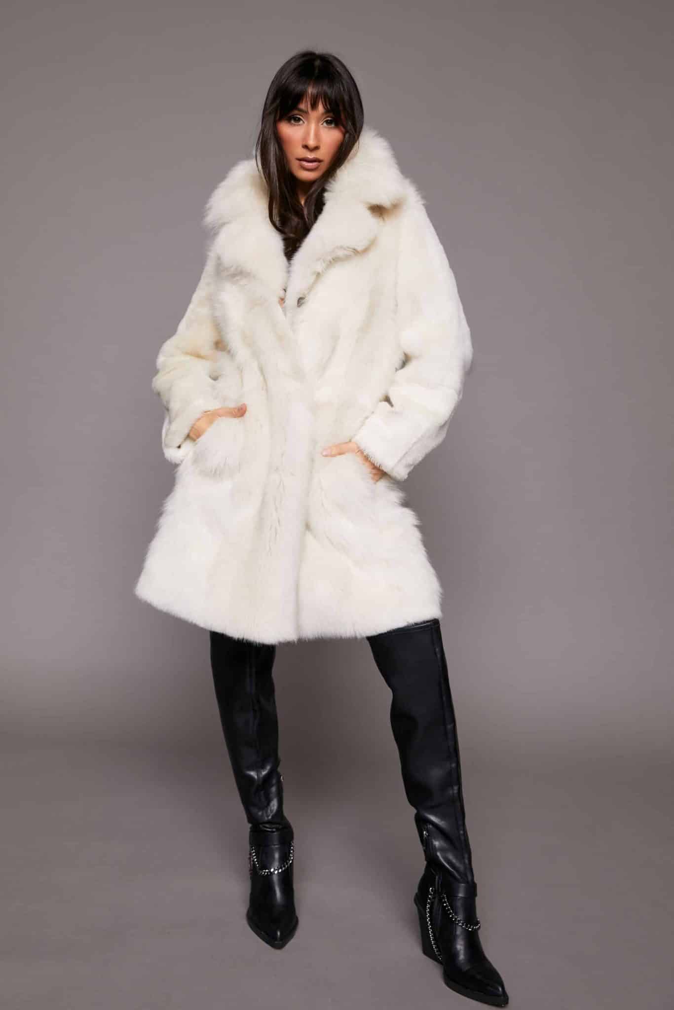 Full body shot of a woman wearing a Toscana shearling coat with the fur side out standing in front of a grey background. The woman's hands are in the coat's pockets.