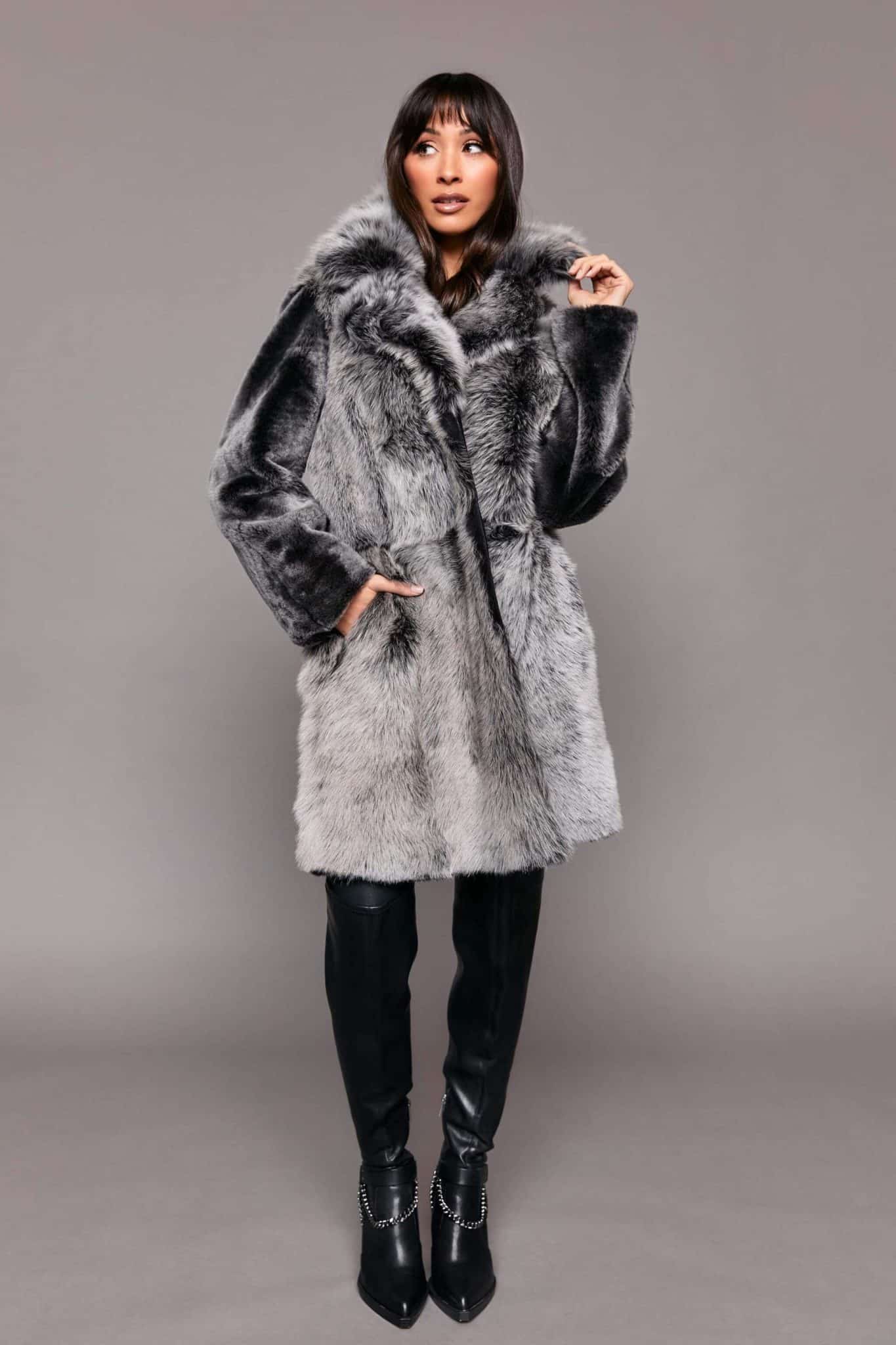 Full body shot of a woman wearing a Toscana shearling coat with the fur side out standing in front of a grey background. The woman's right hand is in the coat's right pocket.