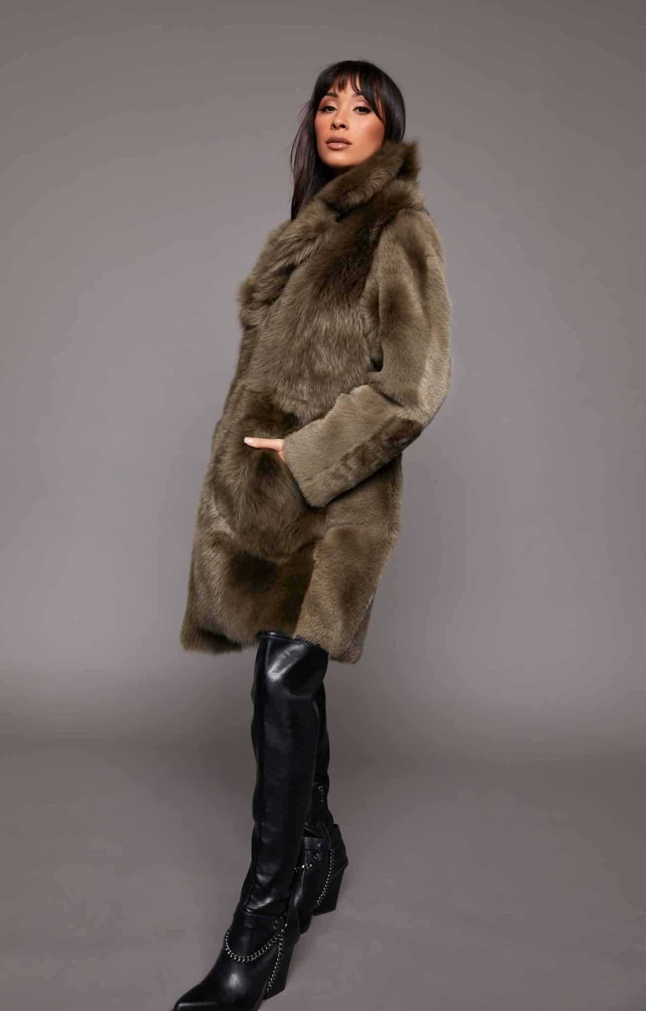 Profile shot of a woman wearing a Toscana shearling coat with the fur side out standing in front of a grey background. The woman's right hand is in the coat's right pocket. Her body is turned towards camera left while her face is pointed towards the camera.