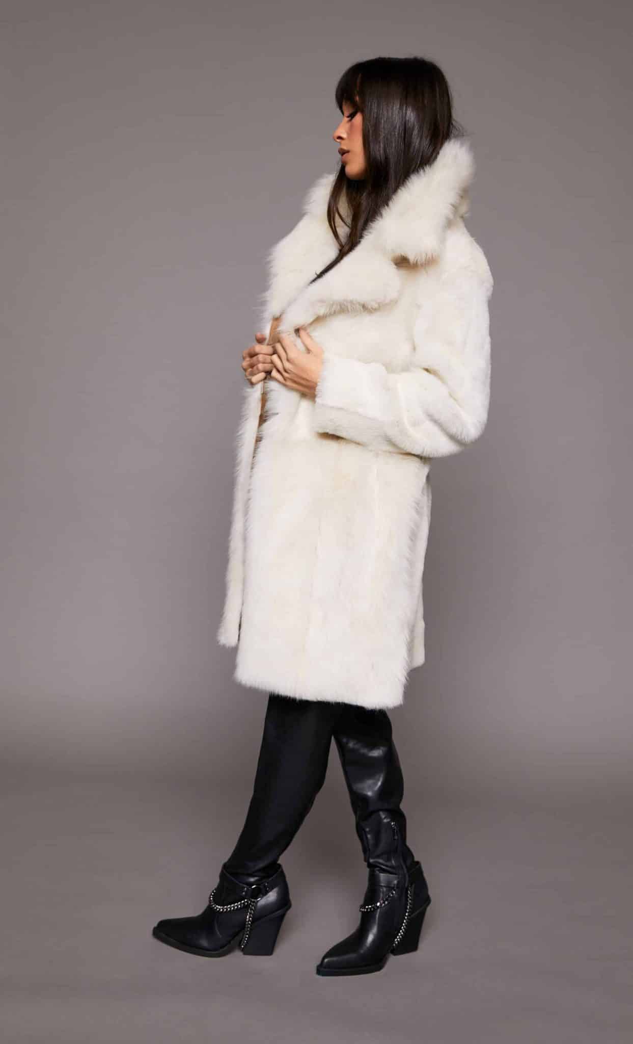 Profile view of a woman wearing a Toscana shearling coat with the fur side out standing in front of a grey background. The woman's hands are clutching the coat's collar while she faces camera left.