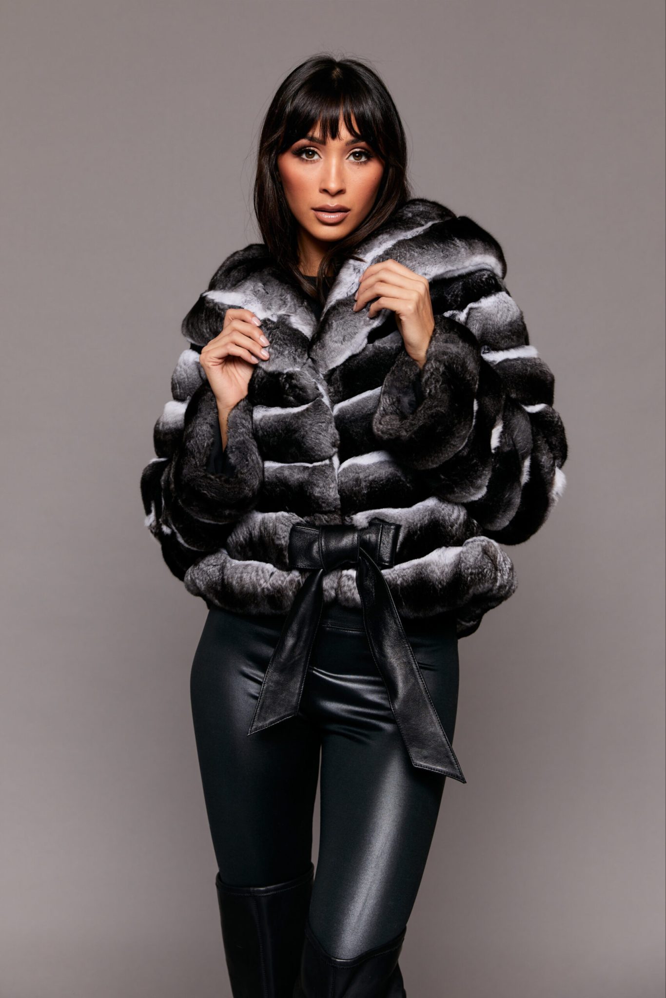 Frontal shot of a woman wearing a black fur coat standing in front of a grey background. The woman's arms are extended upwards so her hands are touching the coat's collar.