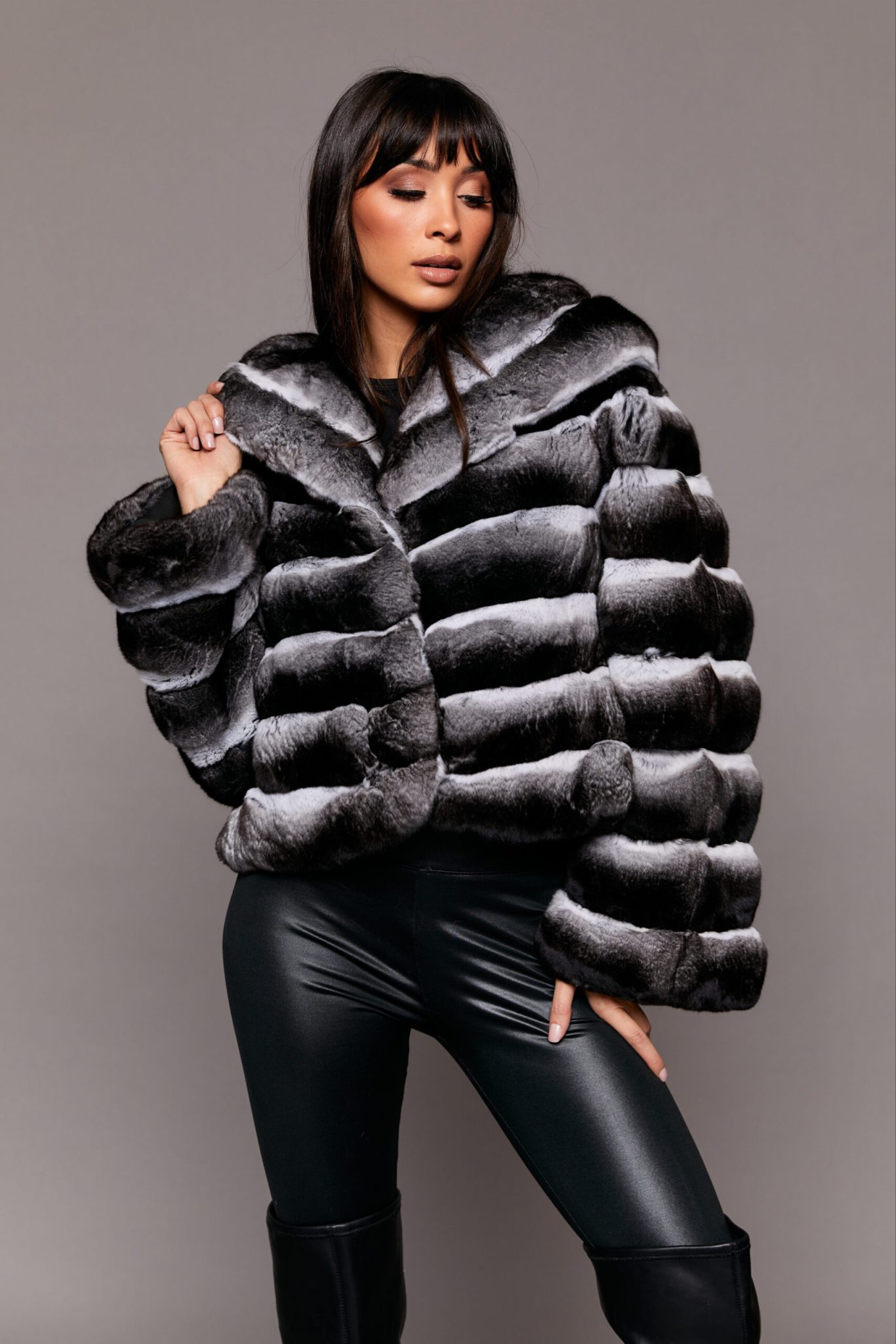 Frontal view of a woman wearing a black fur coat standing in front of a grey background. The woman's left hand is extended upwards to the left side of the jacket's collar while her right hand is touching her right leg.