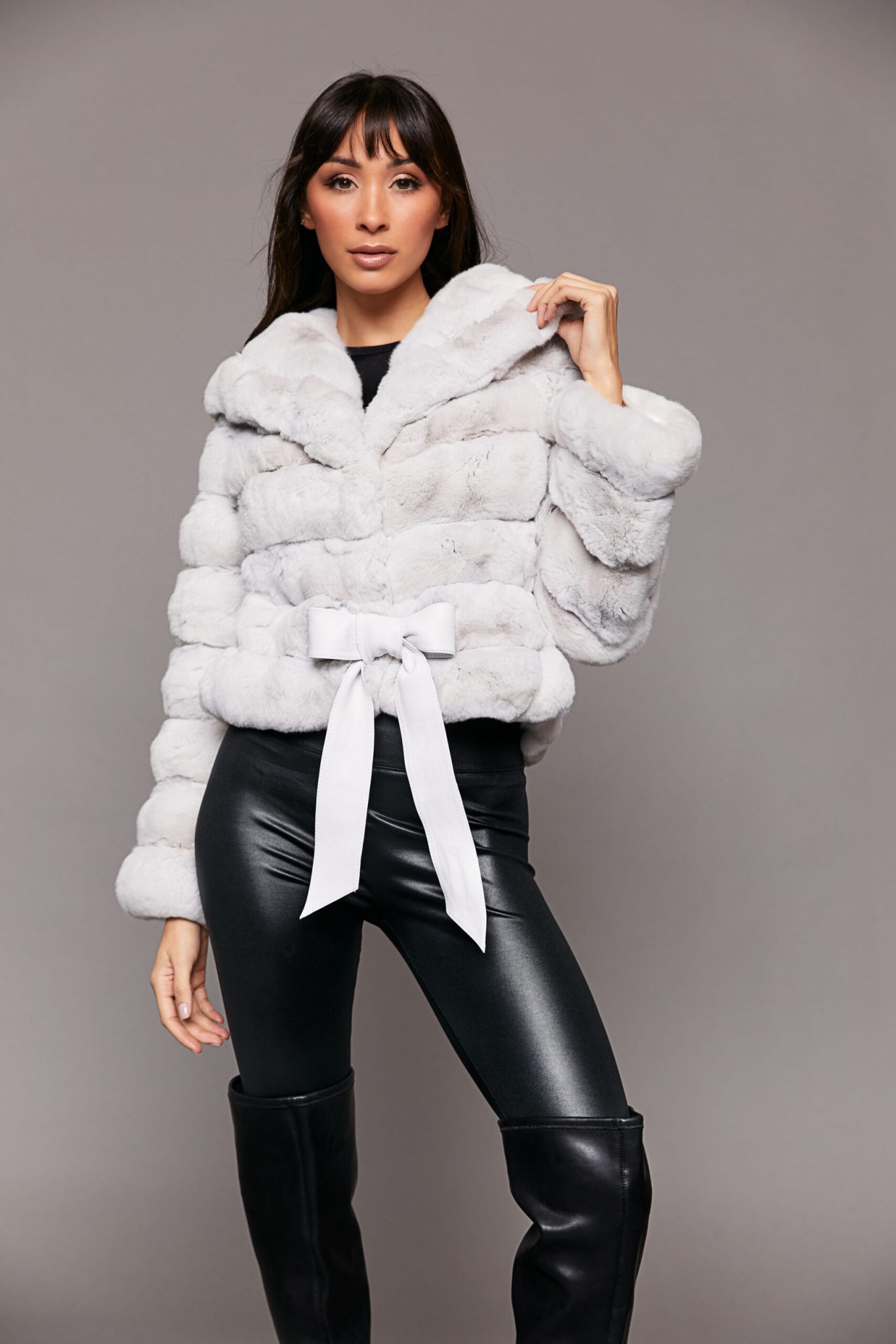 Frontal view of a woman wearing a white Erato Chinchilla Jacket with Shawl Collar with her left hand on the hood standing in front of a seamless backdrop