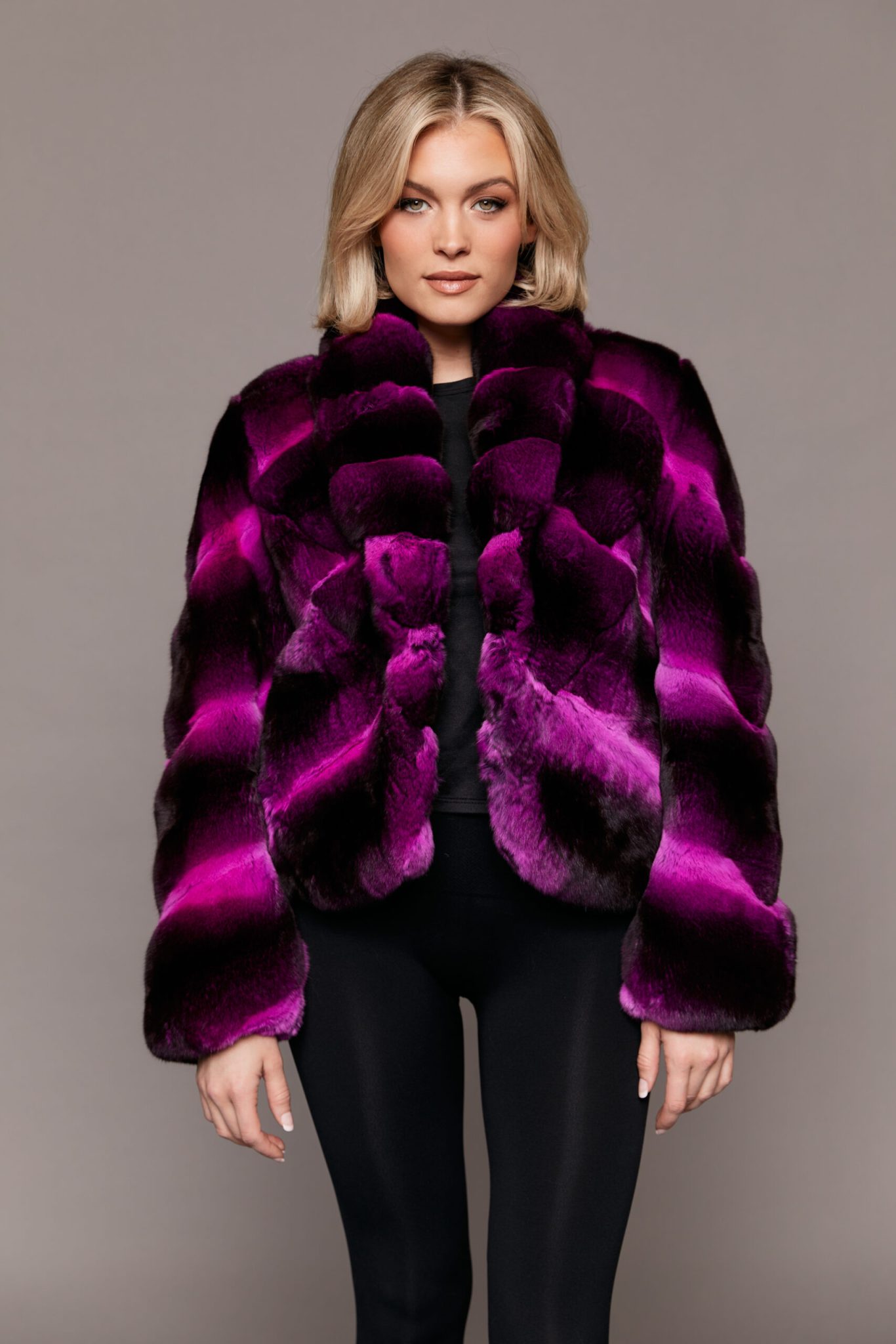 Frontal view of a woman wearing a dyed empress chinchilla fur coat standing in front of a grey background.