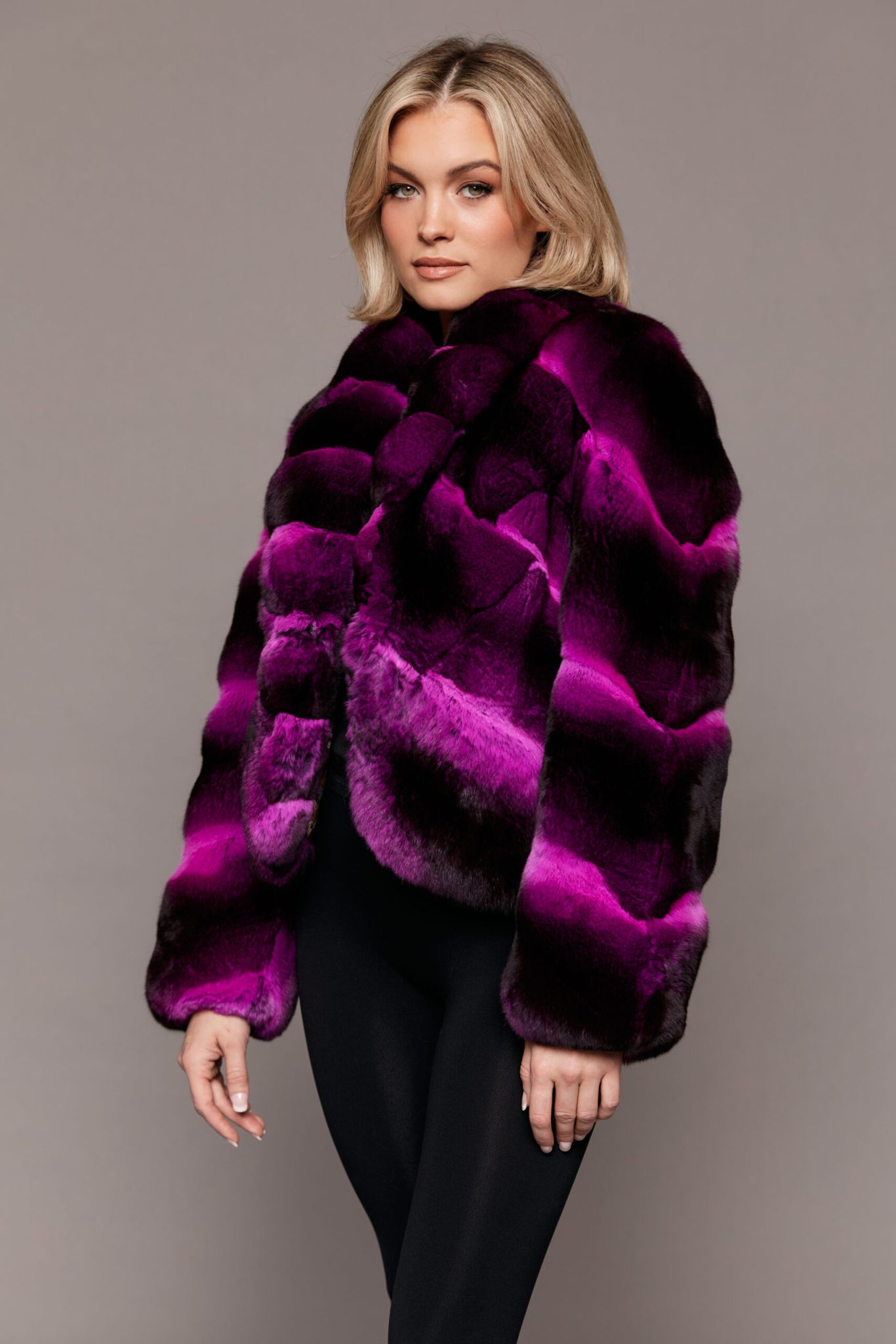 Profile view of a woman wearing a dyed empress chinchilla fur coat standing in front of a grey background. The woman is turned towards camera left while her face is pointed towards the camera.