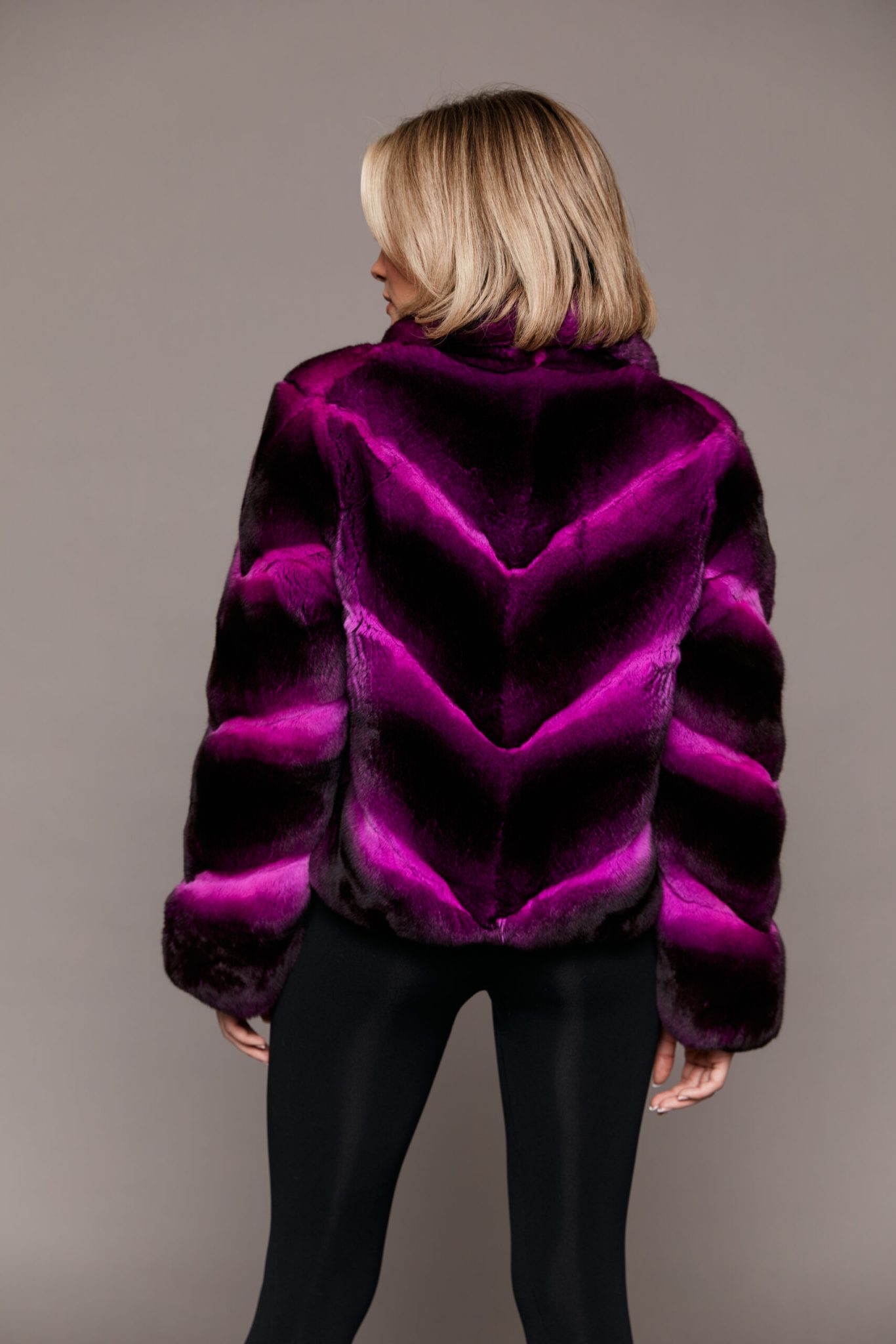 Rear view of a woman wearing a dyed empress chinchilla fur coat facing a grey background.