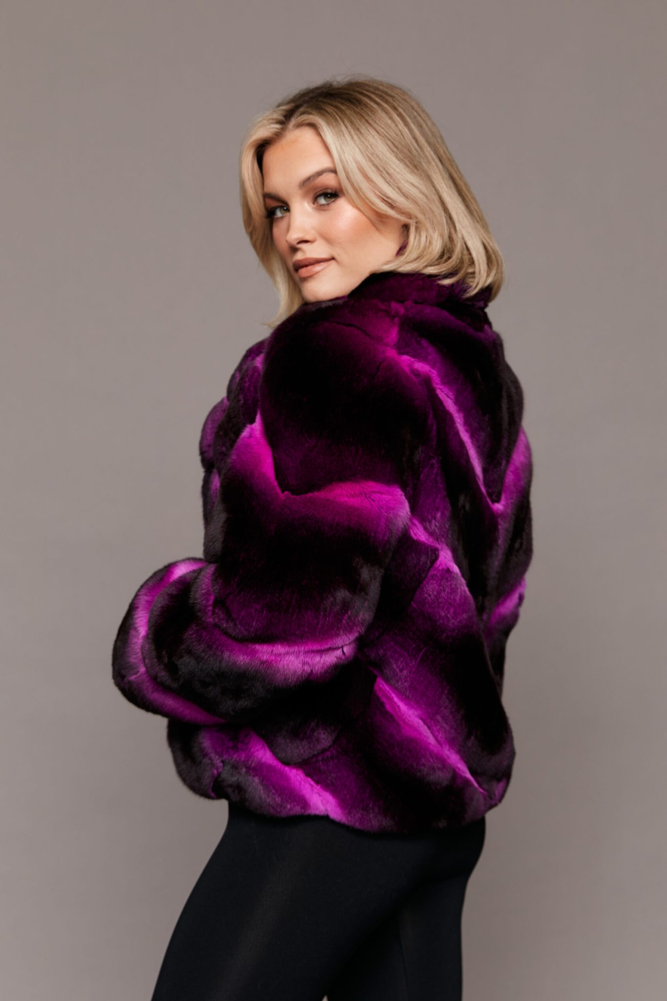 Profile view of a woman wearing a dyed empress chinchilla fur coat standing in front of a grey background. The woman's body is facing camera left while her face is pointed towards the camera.