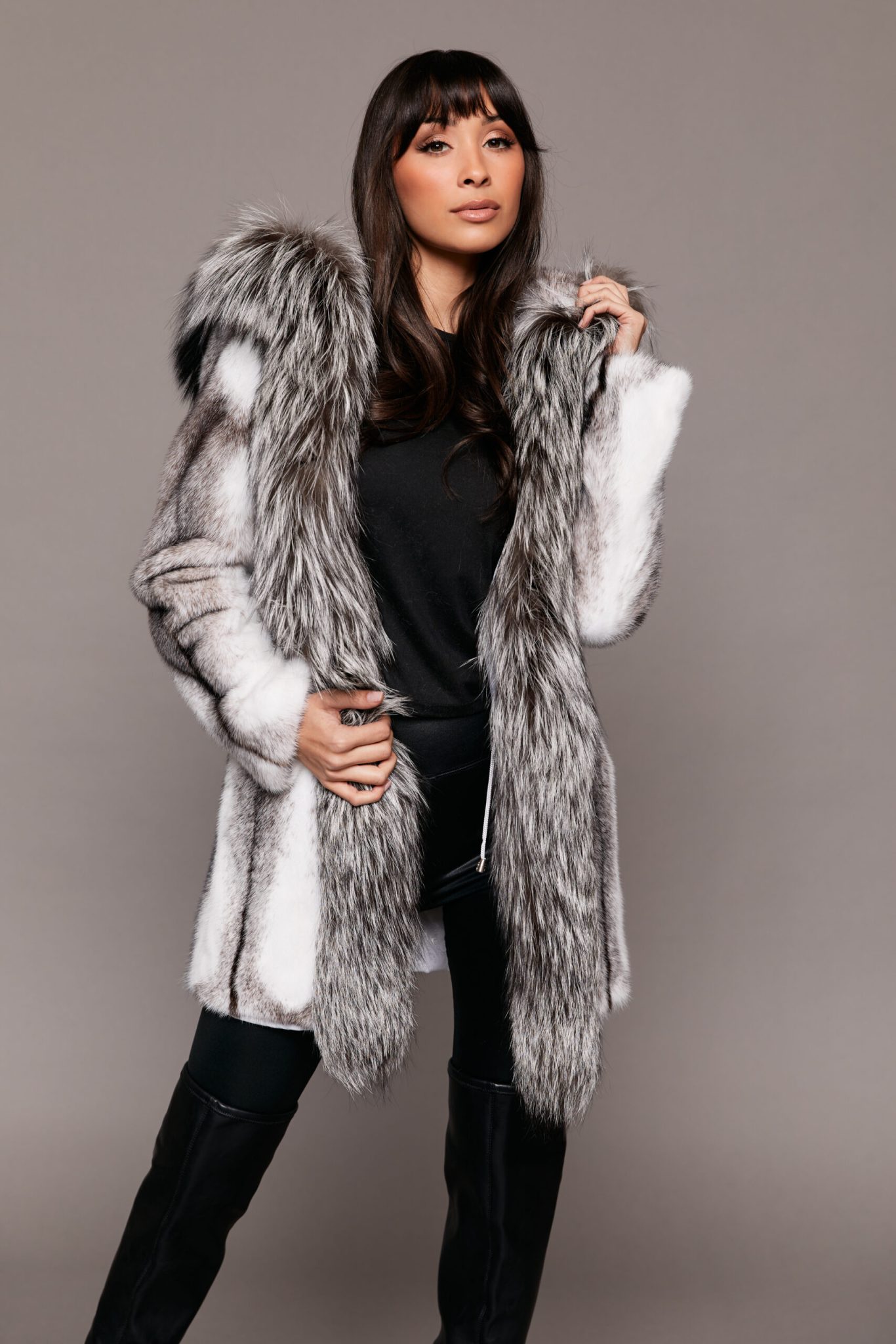 Full body frontal view of a woman wearing a mink fur coat with the hood down standing in front of a grey background.