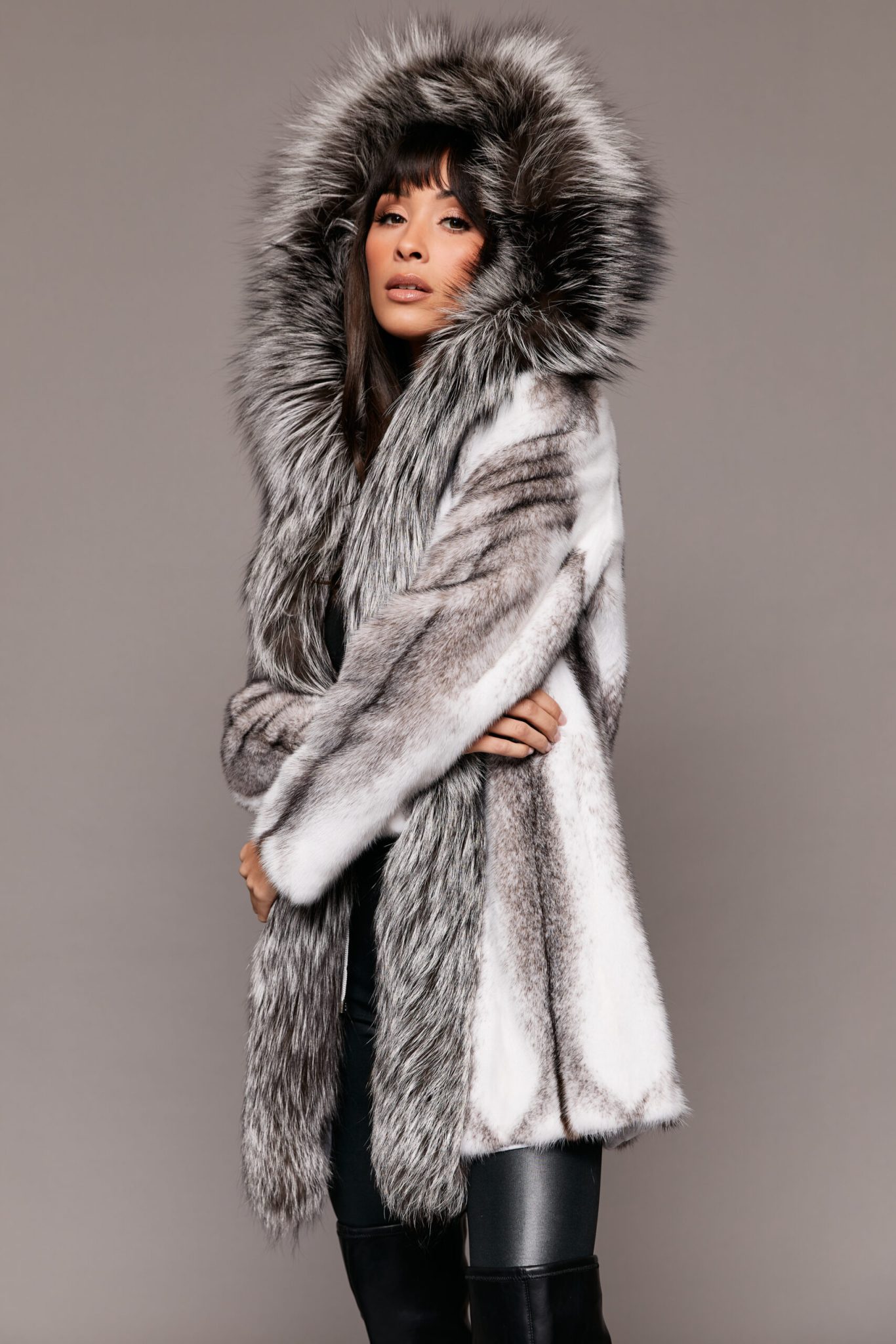 Frontal view of a woman wearing a mink fur coat with the hood up standing in front of a grey background. The woman's arms are crossed.