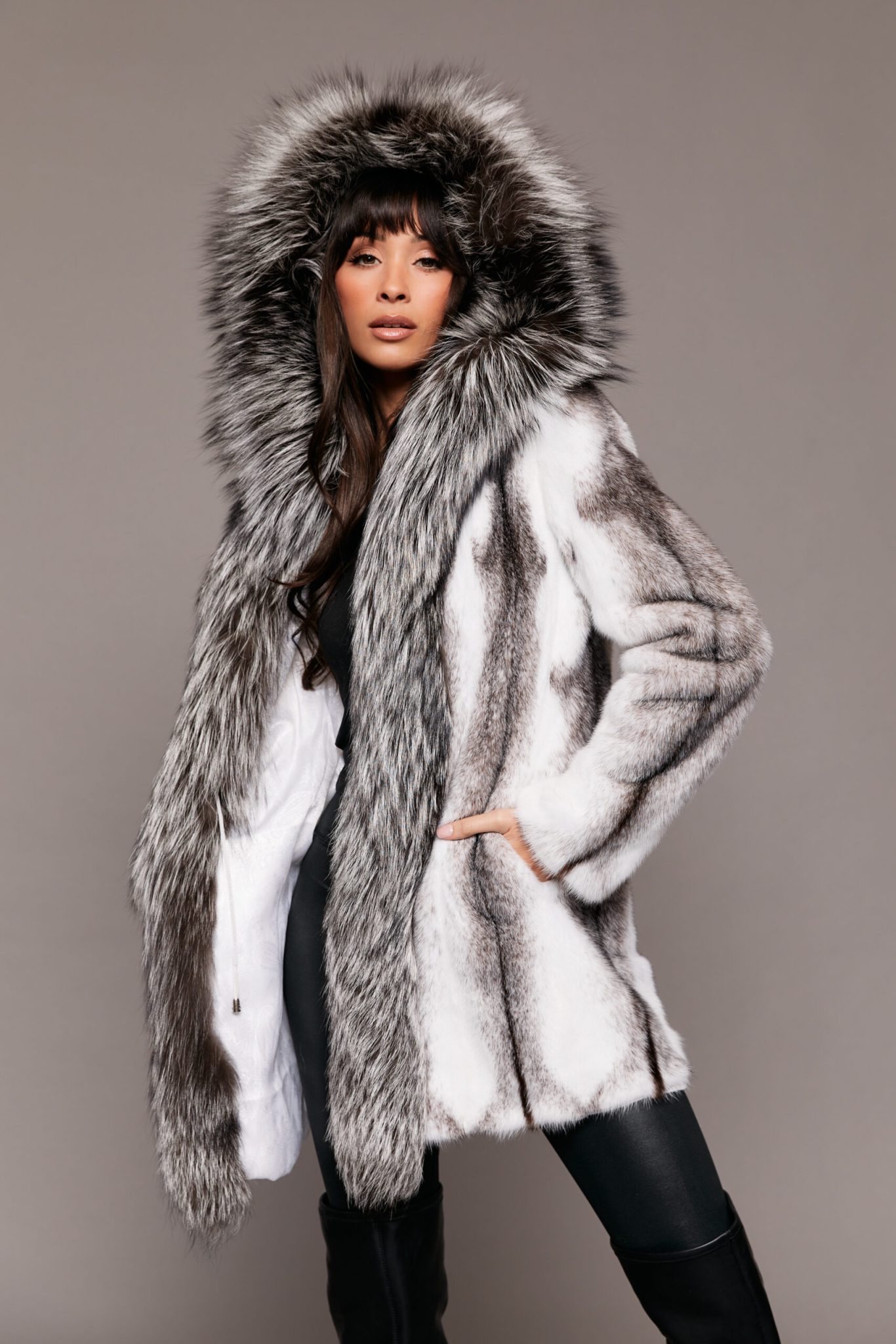 Frontal view of a woman wearing a mink fur coat with the hood up standing in front of a grey background.