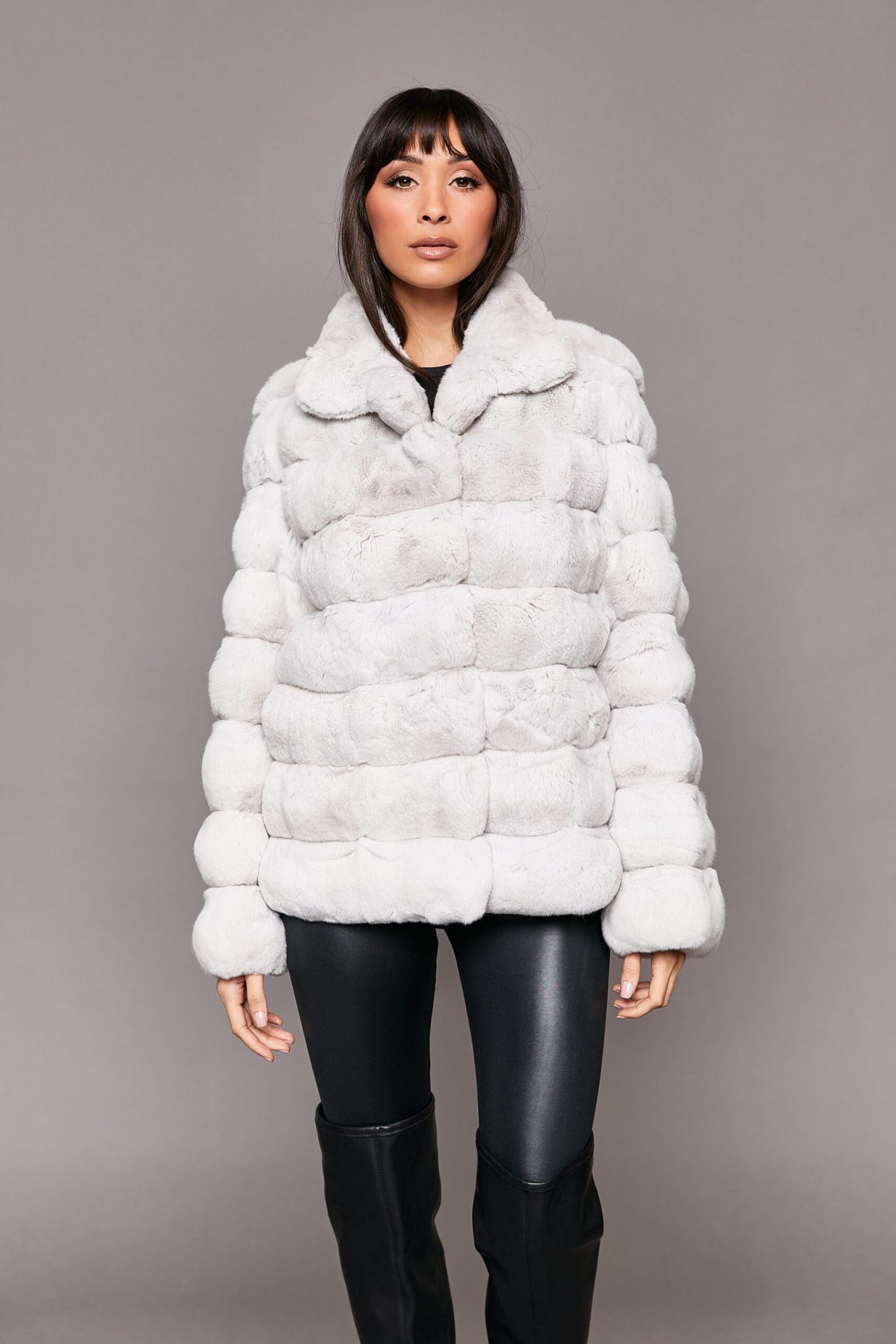 Frontal view of a woman wearing a white genuine chinchilla coat with her arms to her side and one leg slightly forward standing in front of a seamless backdrop
