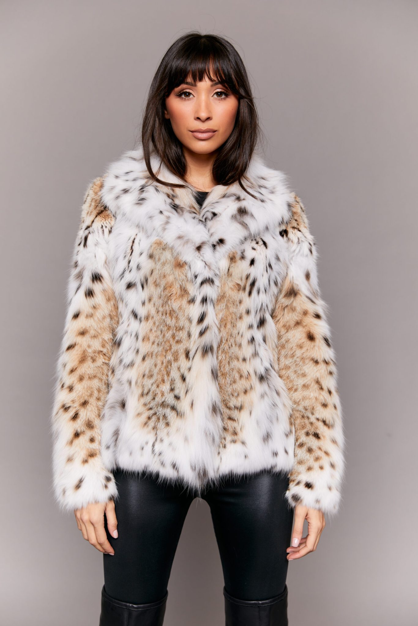 Frontal view of a woman wearing a lynx fur jacket standing in front of a grey background. The woman's hands are at her sides.