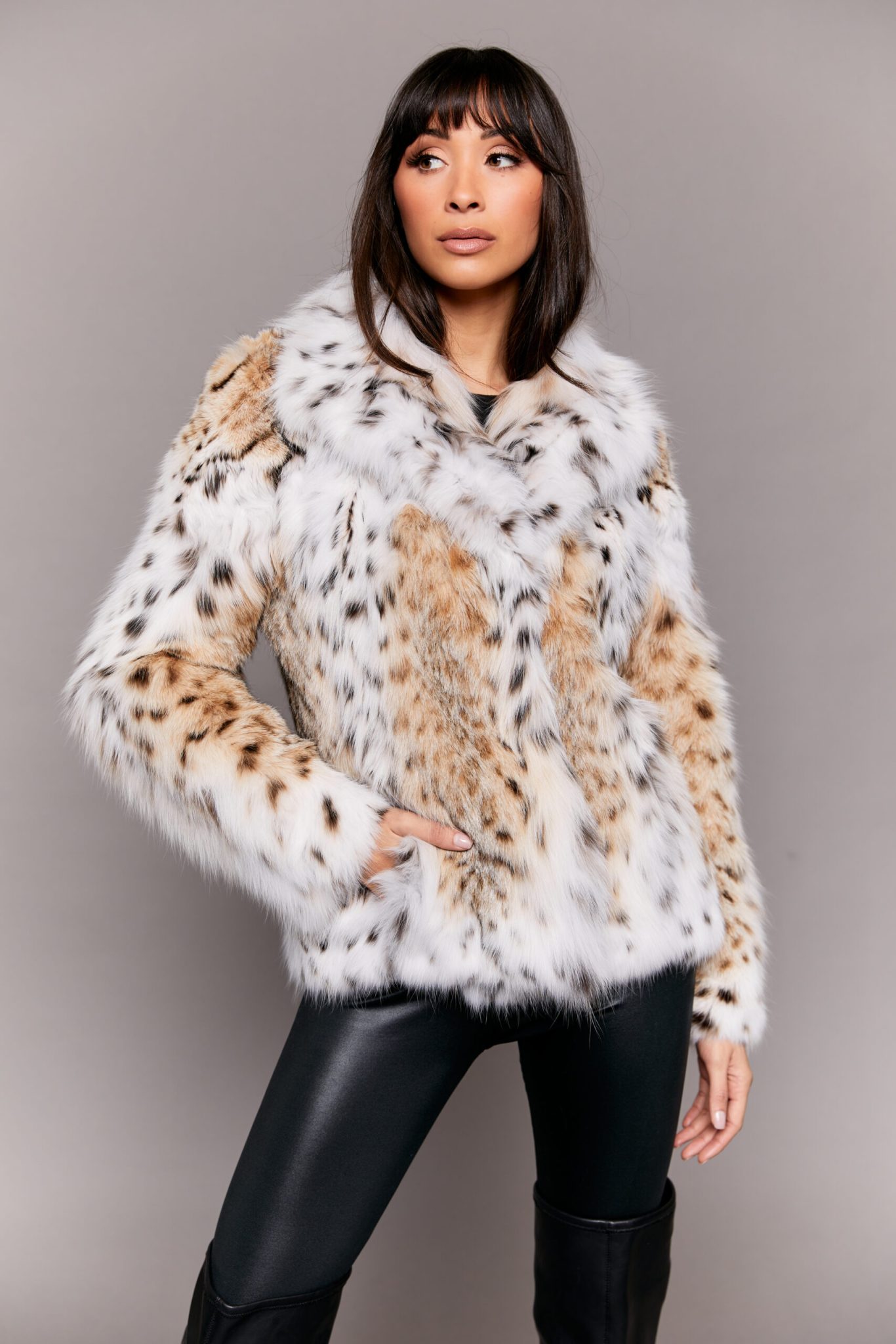 Frontal view of a woman wearing a lynx fur jacket standing in front of a grey background. The woman has her right hand in the jacket's right pocket.