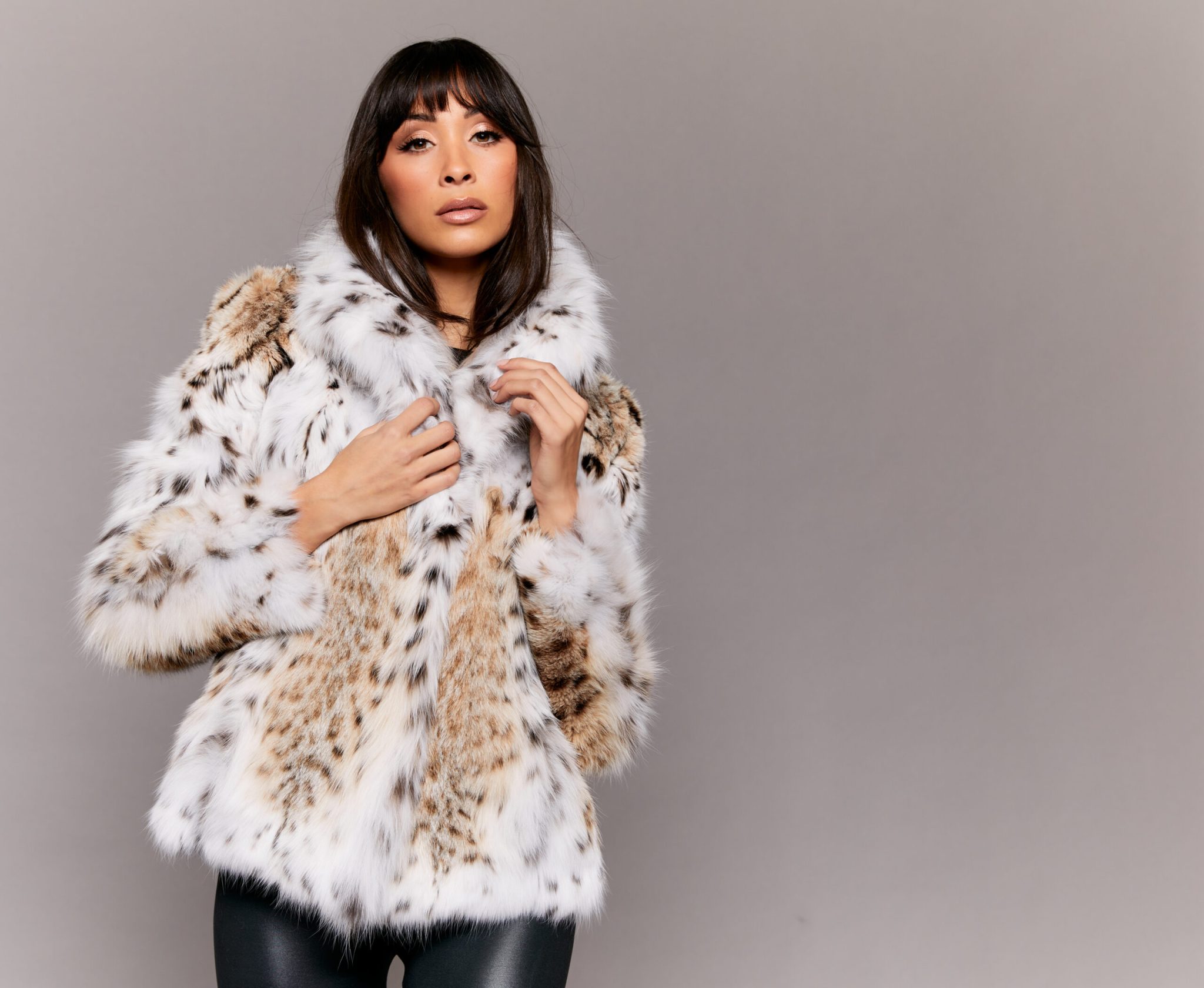 Frontal view of a woman wearing a lynx fur jacket standing in front of a grey background. The woman's hands are holding the lower portion of the hood.