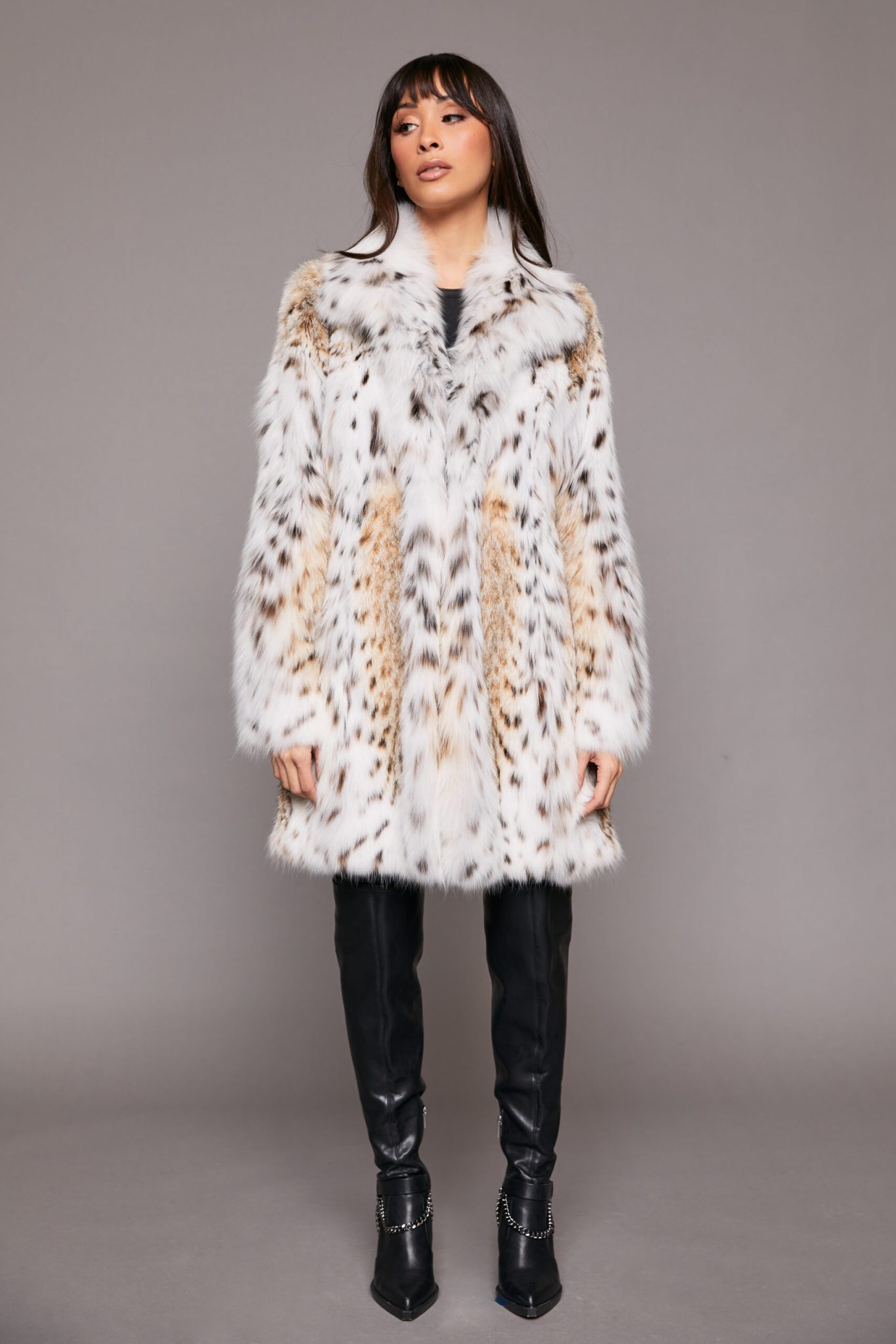 Frontal view of a woman with dark brown hair wearing a lynx fur jacket standing in front of a grey background.
