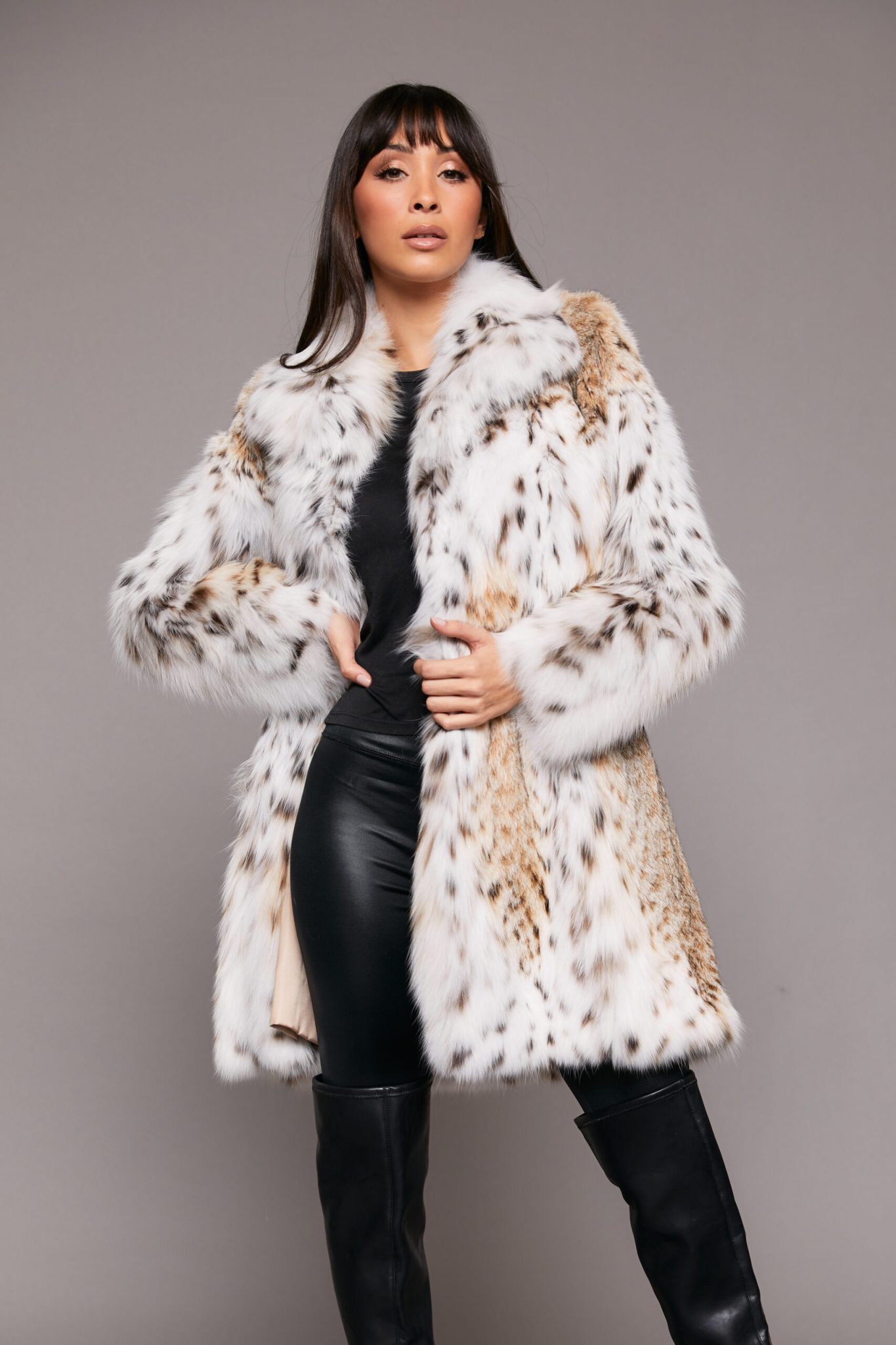 Frontal view of a woman with long brown hair wearing a lynx fur jacket and her right arm on her right hip standing in front of a grey background.