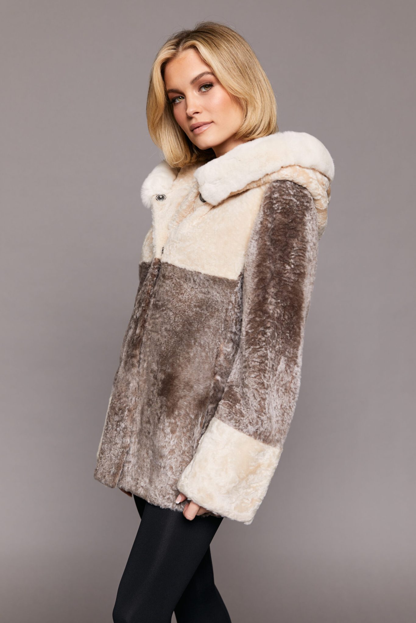 Profile view of a woman wearing a reversible Astrakhan coat with the hood down standing in front of a grey background. The woman's body is turned to camera left but her face is turned towards the camera.