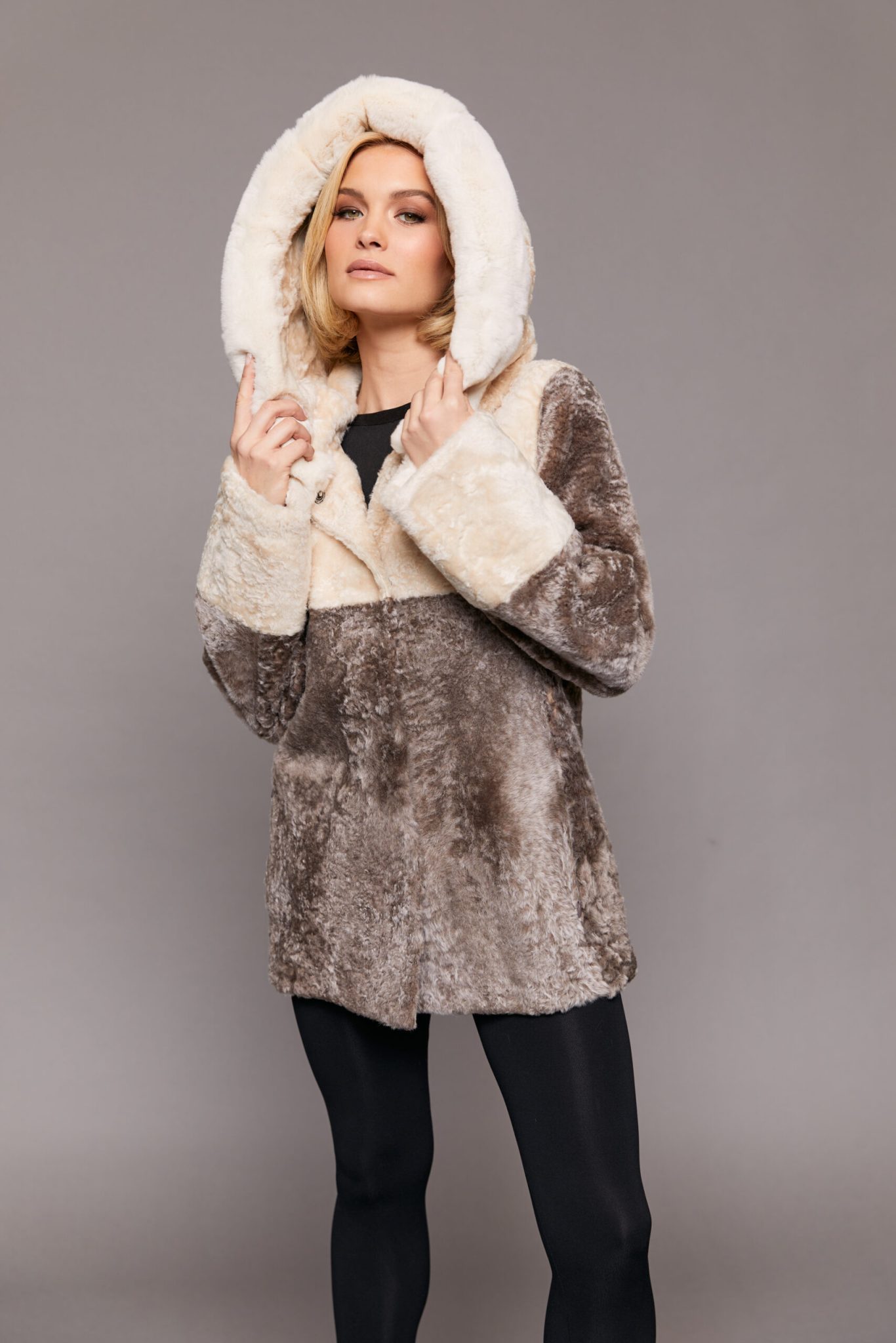 Frontal view of a woman wearing a reversible Astrakhan coat with the hood up standing in front of a grey background. The woman's hands are clutching the hood
