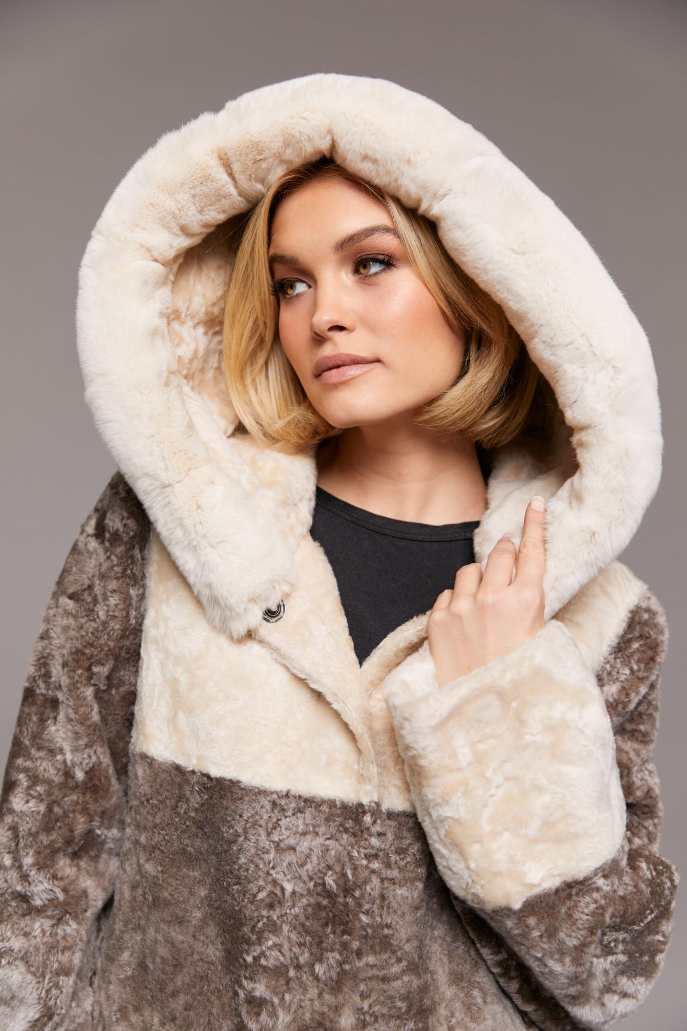 Close-up frontal view of a woman wearing a reversible Astrakhan coat with the hood up standing in front of a grey background.
