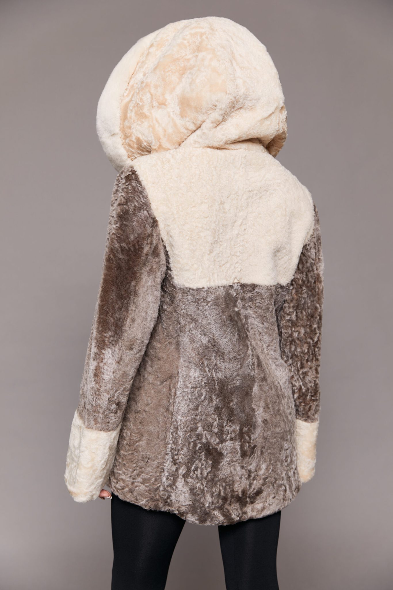 Rear view of a woman wearing a reversible Astrakhan coat with the hood up facing a grey background.