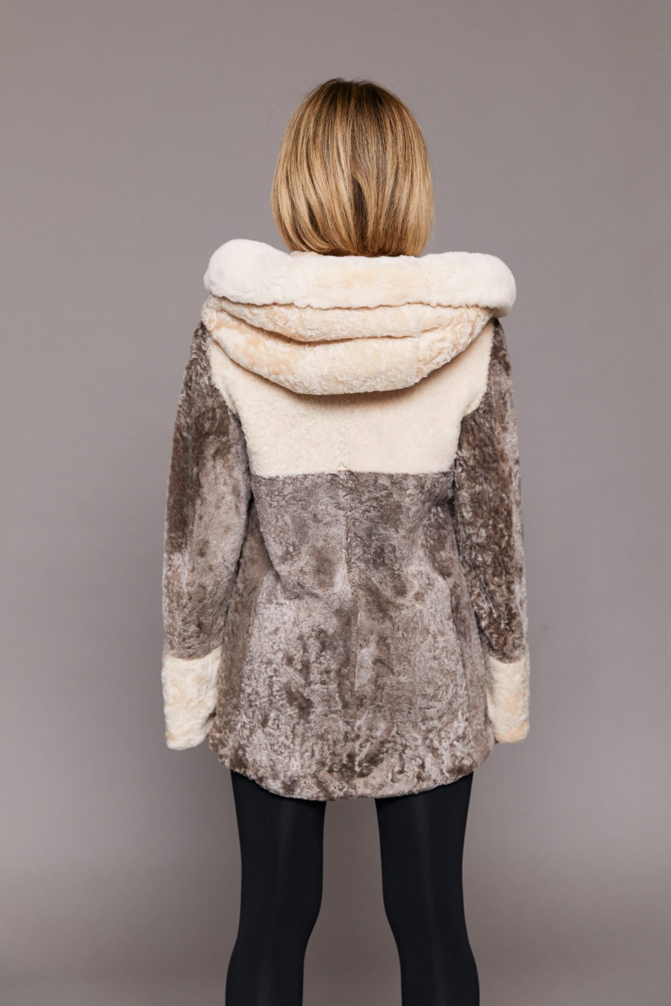 Rear view of a woman wearing a reversible Astrakhan coat with the hood down facing a grey background.