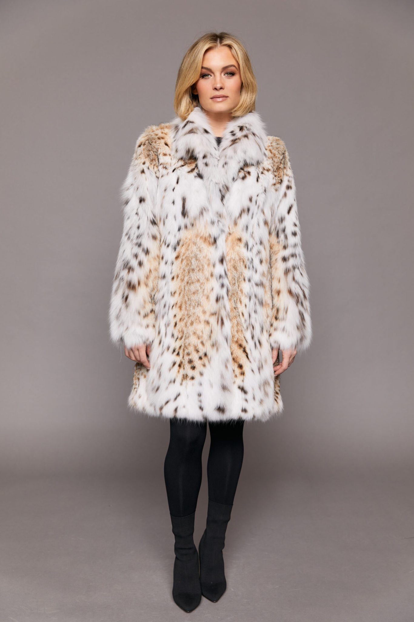 Frontal view of a woman wearing a lynx fur jacket facing the camera in front of a grey background, her arms are at her sides and the jacket is closed