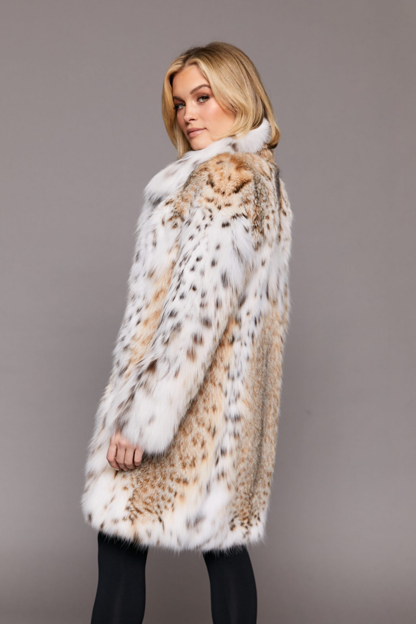 Side view of a woman wearing a lynx fur jacket with her arms at her sides, her head is turned towards the camera and she is standing in front of a grey background.