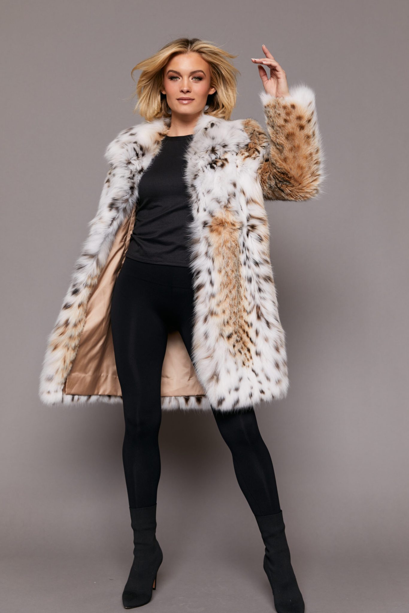 Frontal view of a woman wearing a lynx fur jacket with her left arm raised in the air. The jacket is open and the hood is down. The woman is standing in front of a grey background.