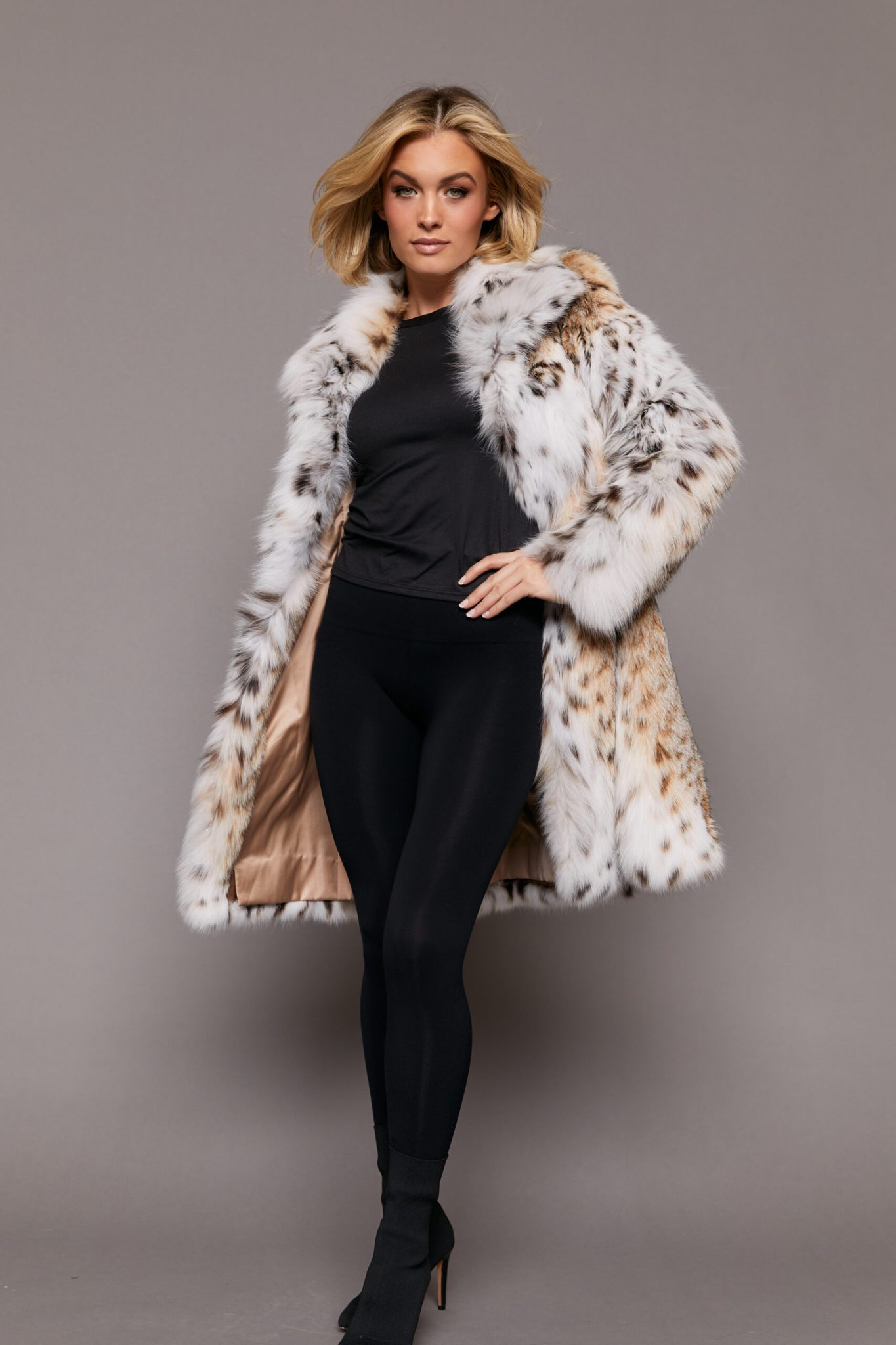 Frontal view of a woman wearing a lynx fur jacket with her left arm on her left hip. The jacket is open and the hood is down.