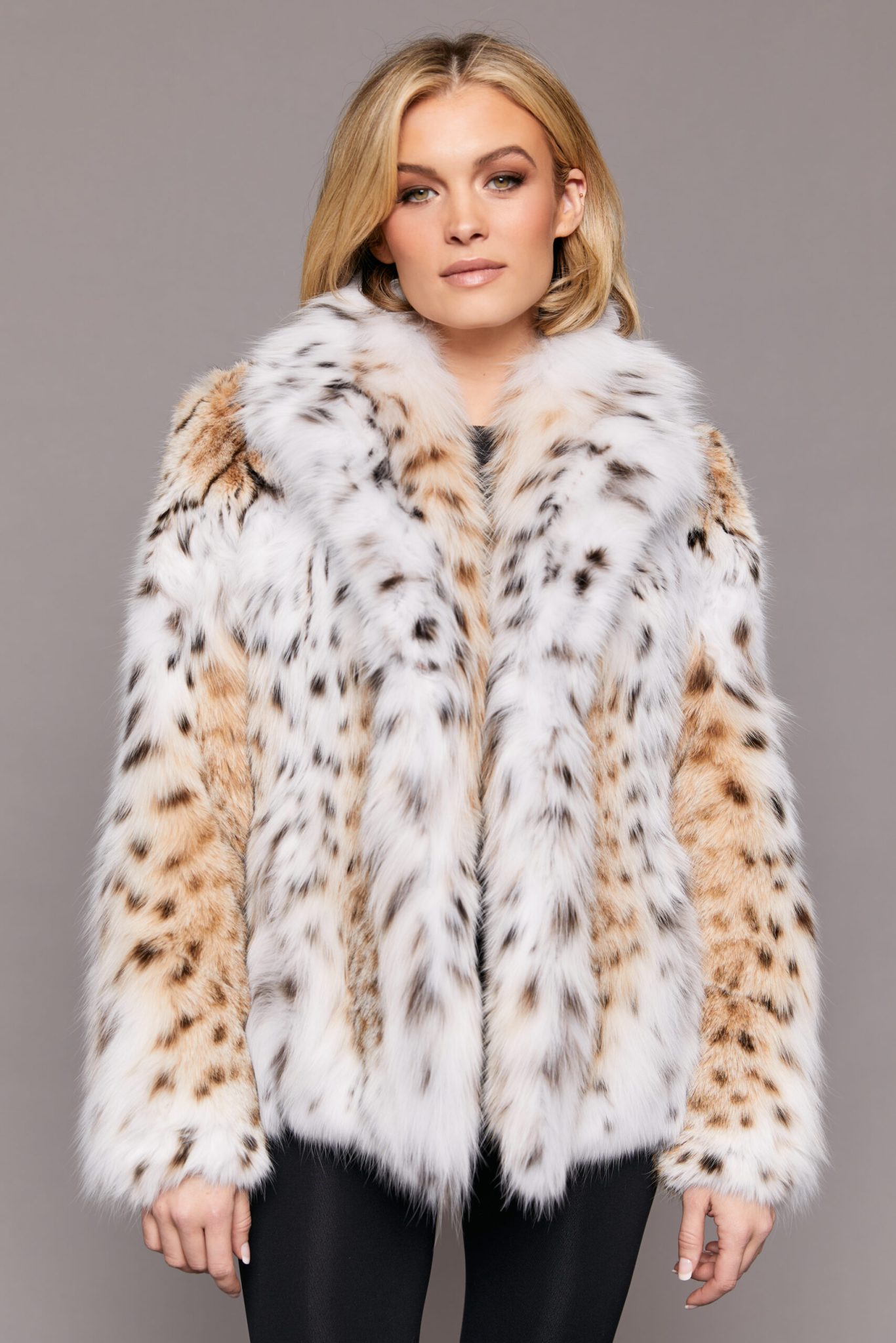 Medium frontal close-up of a woman wearing a lynx fur jacket. Her arms are at her sides and she is standing in front of a grey background.