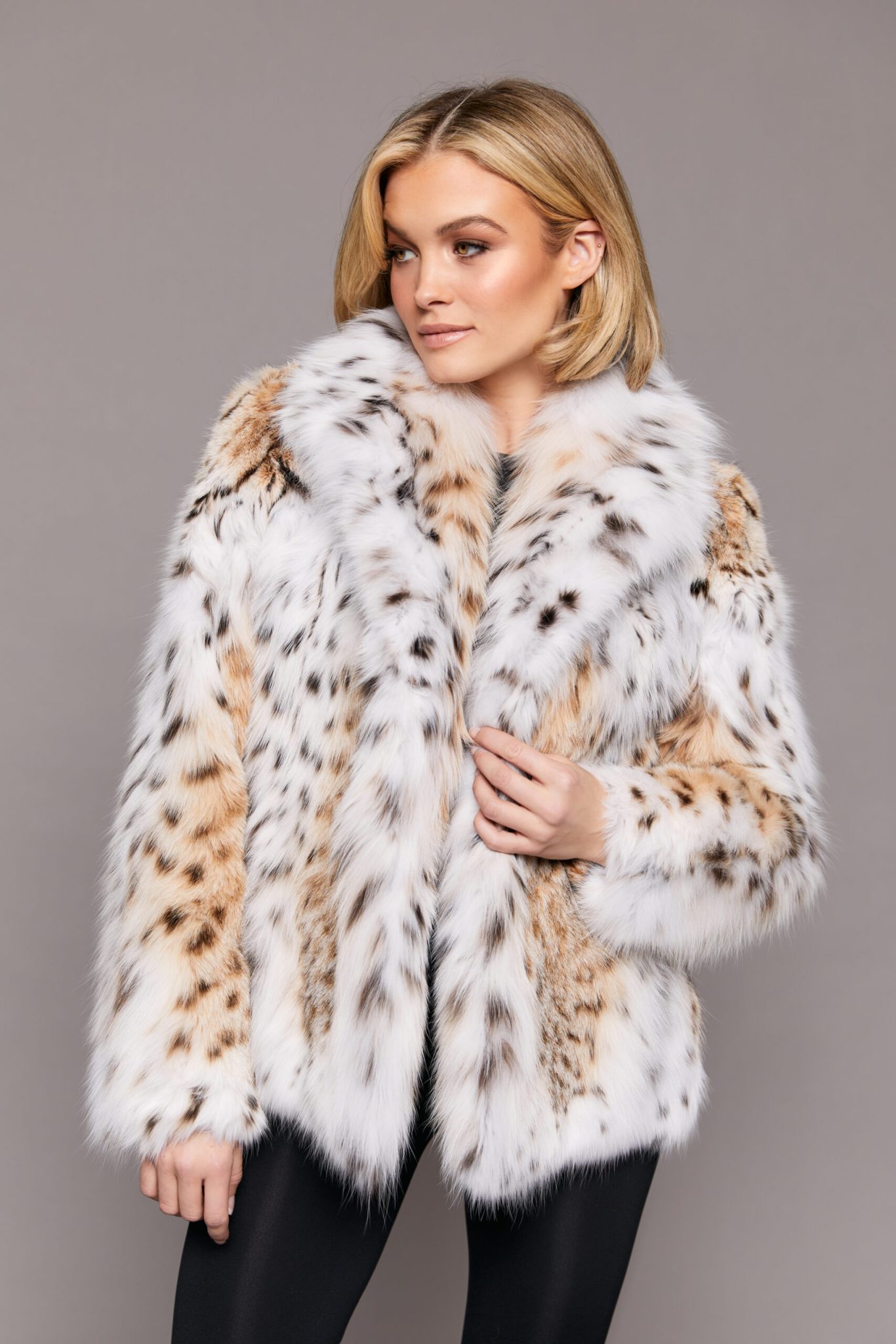 Frontal view of a blonde woman wearing a lynx fur jacket standing in front of a grey background.