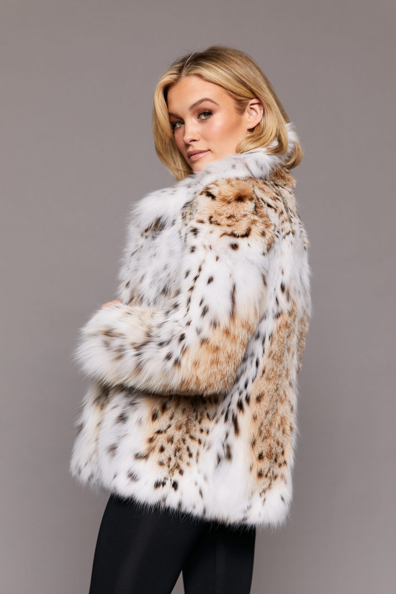 Profile view of a woman wearing a lynx fur jacket standing in front of a grey background. The woman's body is facing camera left while her face is turned towards the camera.