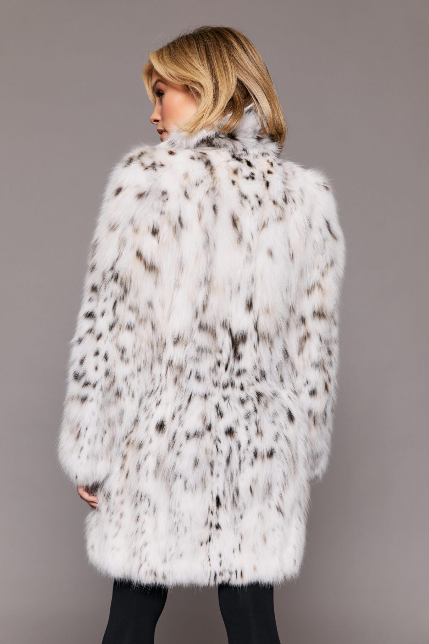 Rear view of a woman wearing a white fur coat turned towards a grey background. The woman's head is turned to the left of frame.