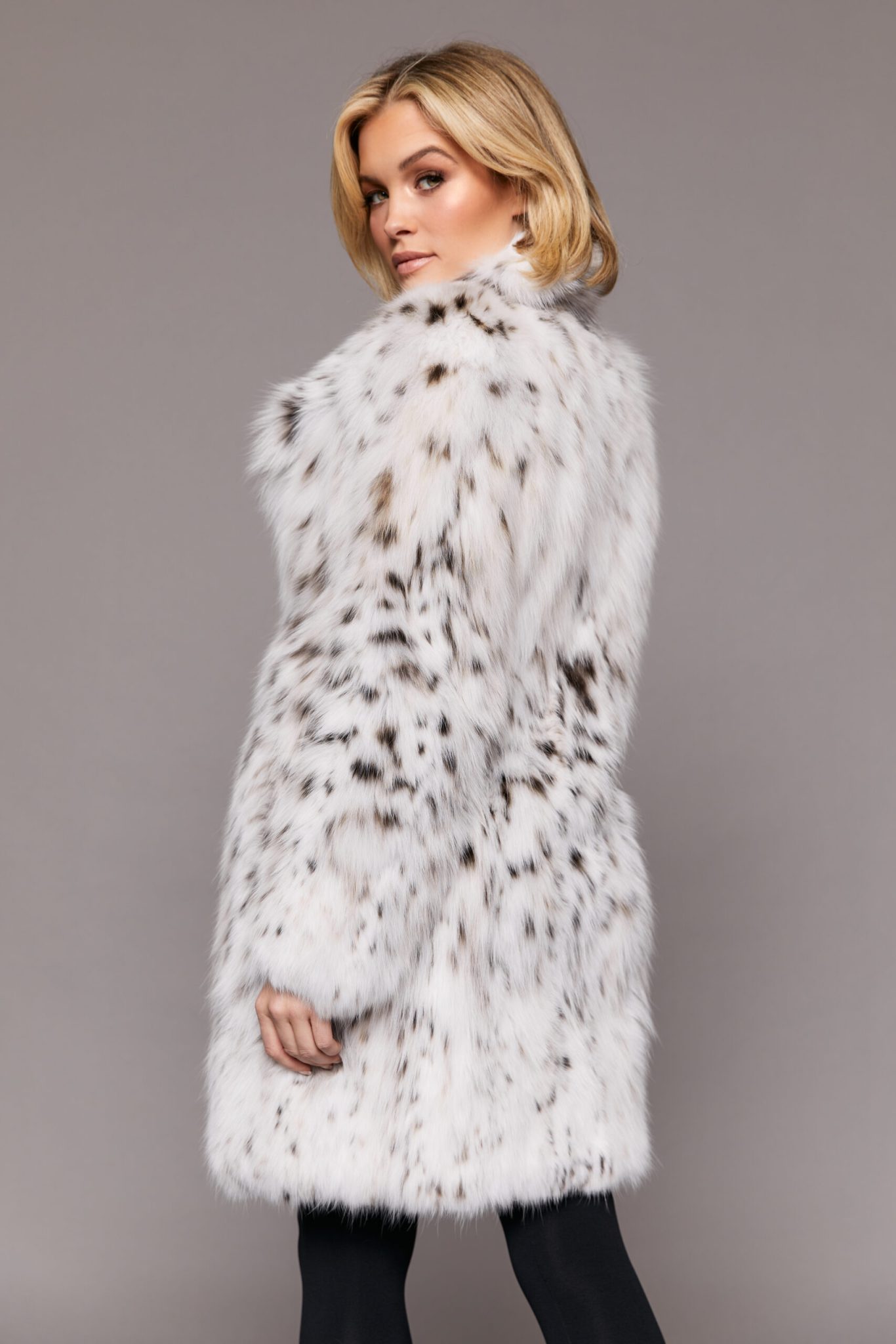 Profile view of a woman wearing a white fur coat standing in front of a grey background. The woman's left hand is closes to the camera and lowered to her side while the woman looks past the camera lens.