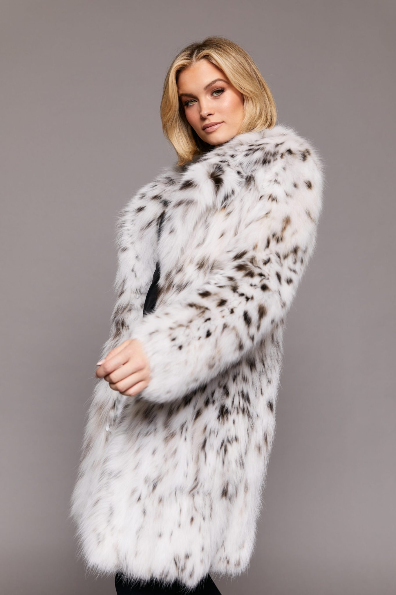 Frontal view of a woman wearing a white fur coat standing in front of a grey background. The woman's left hand is frozen in movement as it swings in front of her body.