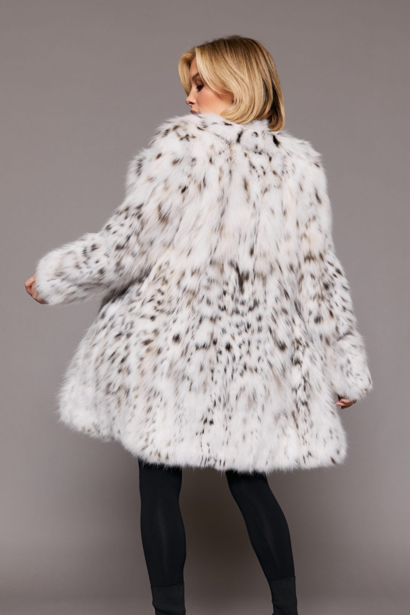Rear view of a woman wearing a white fur coat standing in front of a grey background. The woman's left hand is frozen in a movement to the left as it extends away from her body.