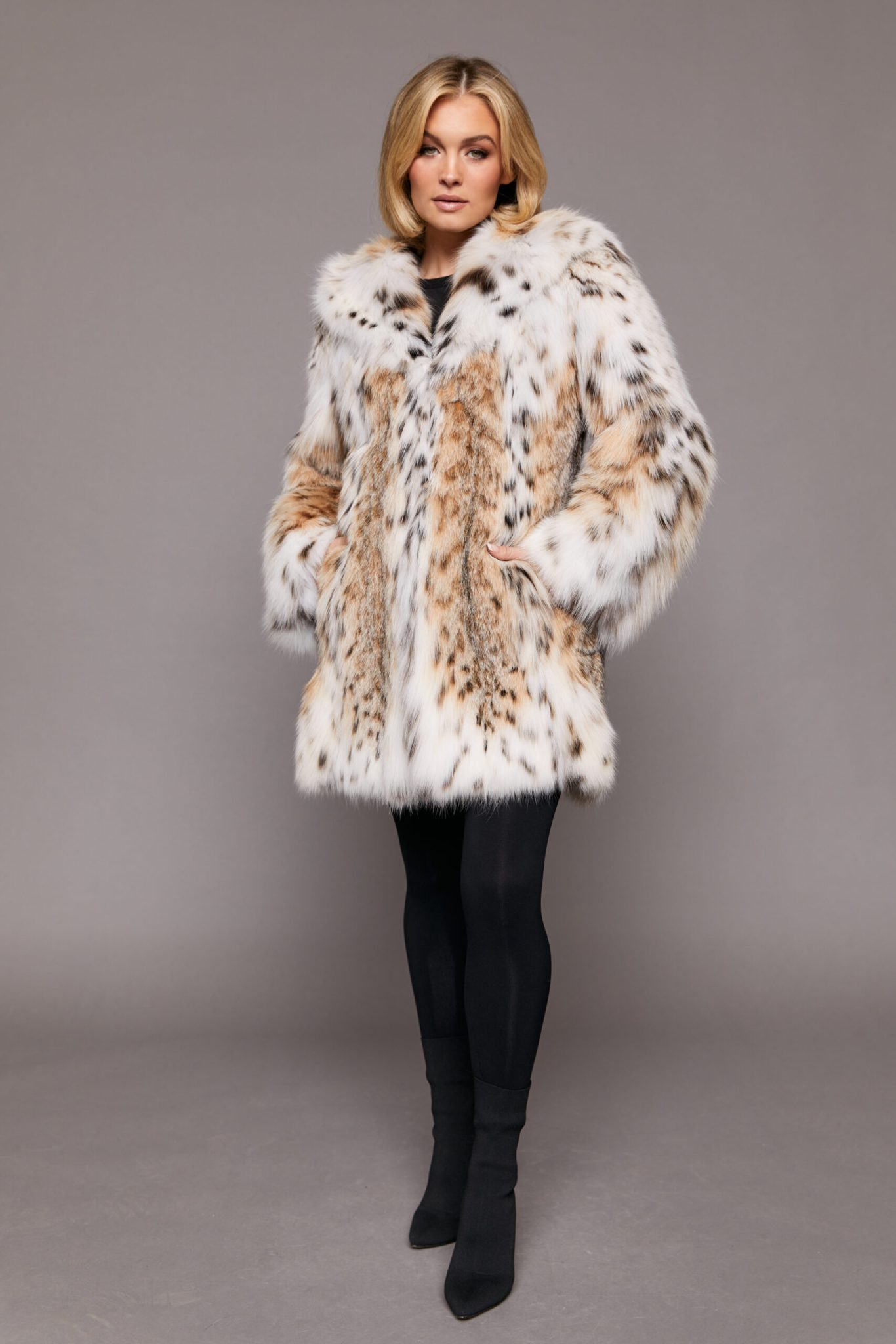 Frontal full body view of a woman wearing a fur coat standing in front of a grey background. The woman's hands are in the coat's front pockets.