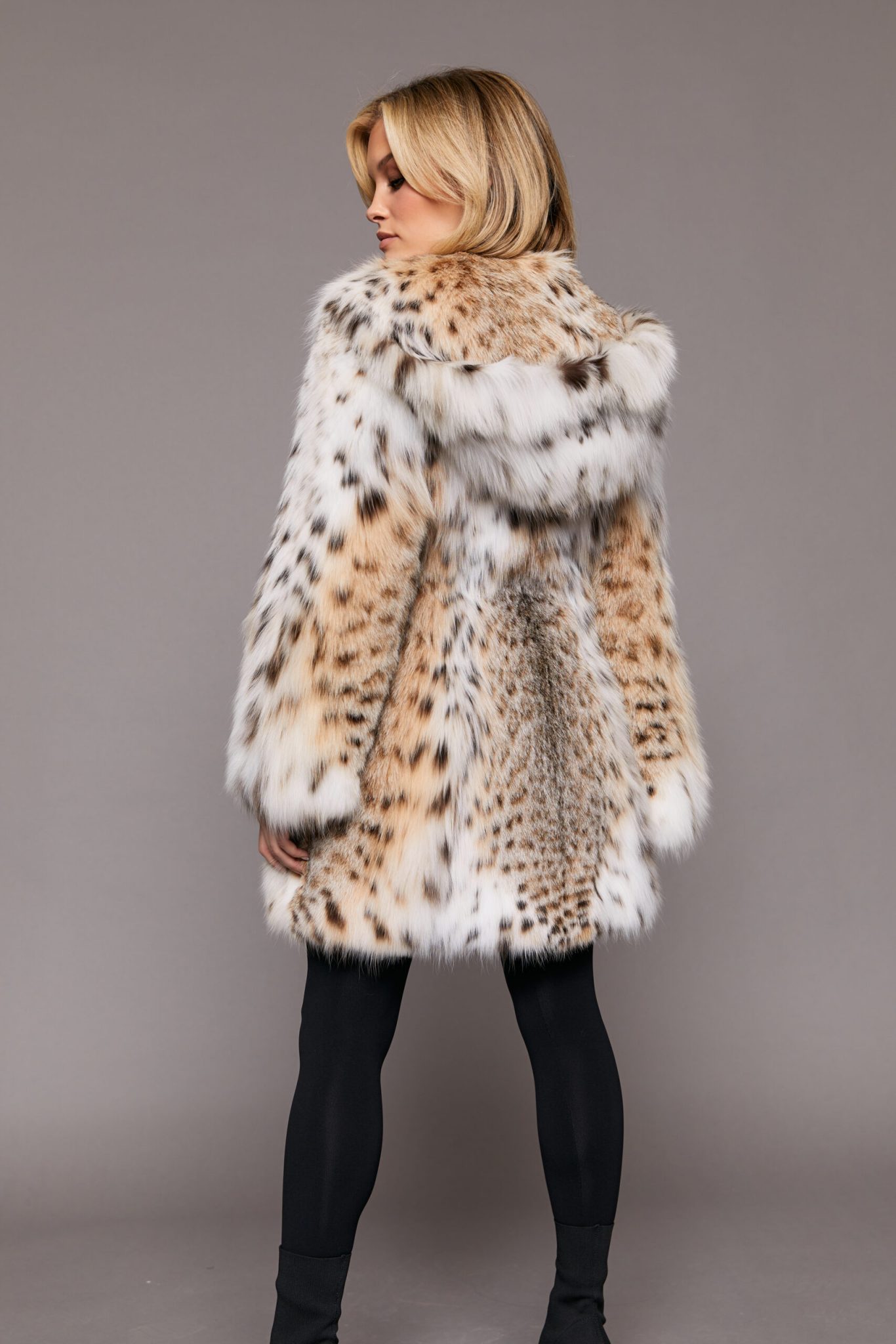 Rear view of a woman wearing a fur coat turned towards a grey background. The woman is looking to the left of frame and her arms are at her sides.