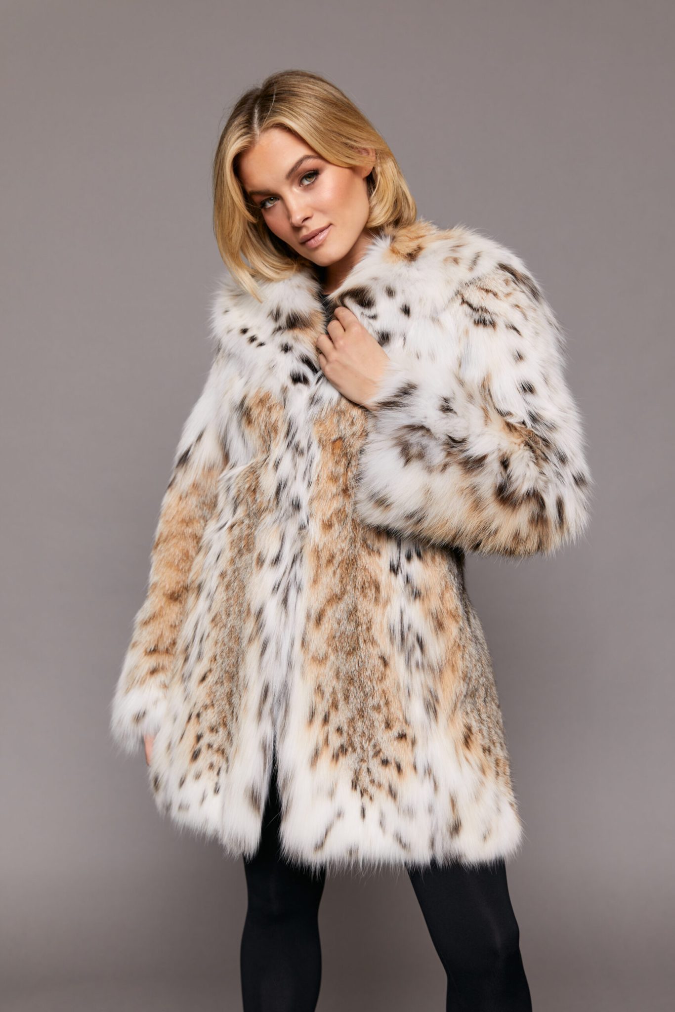 Frontal view of a woman wearing a fur coat standing in front of a grey background. The woman's left hand is touching the collar and her right hand is at her side.
