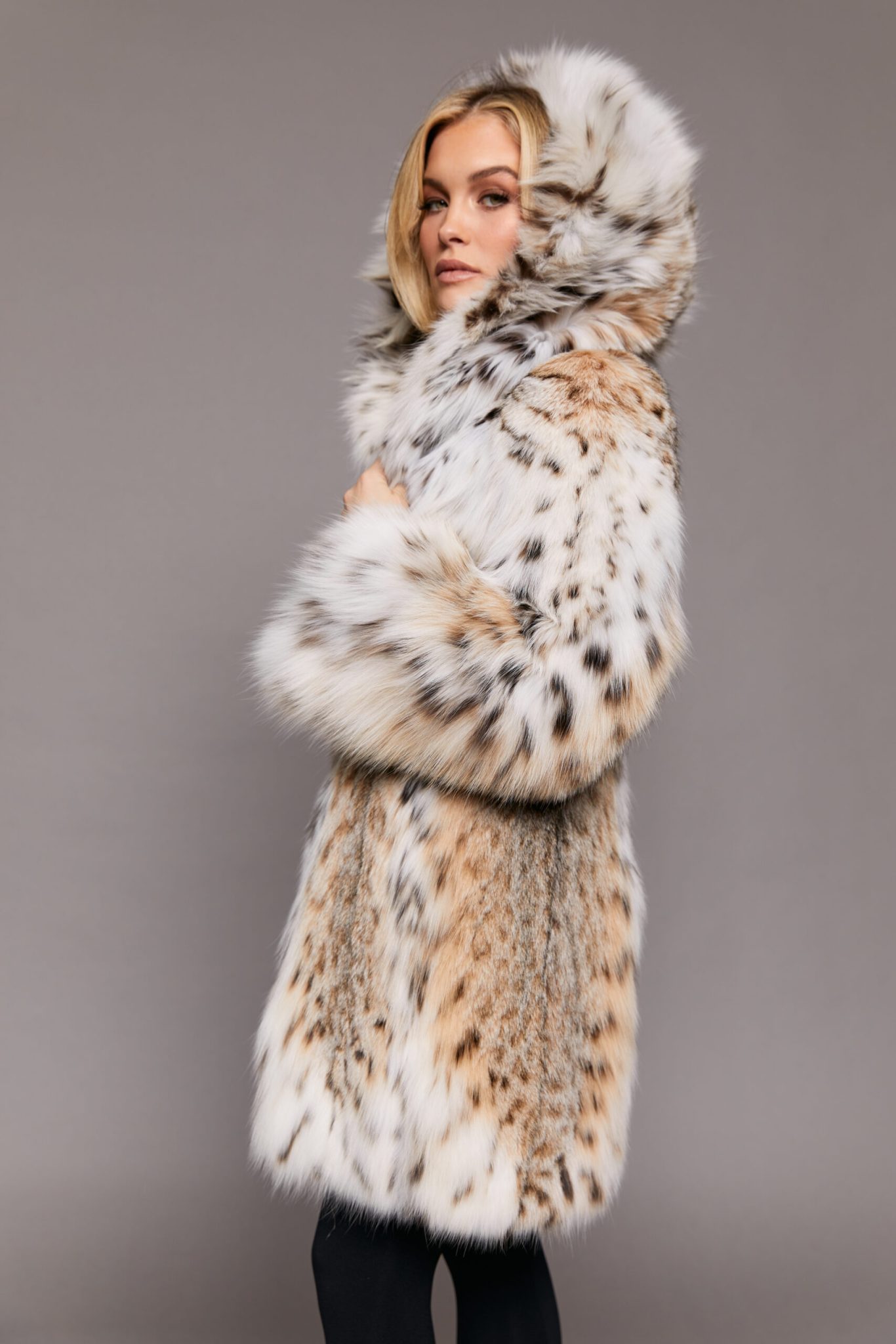 Profile view of a woman wearing a fur coat with the hood up standing in front of a grey background.