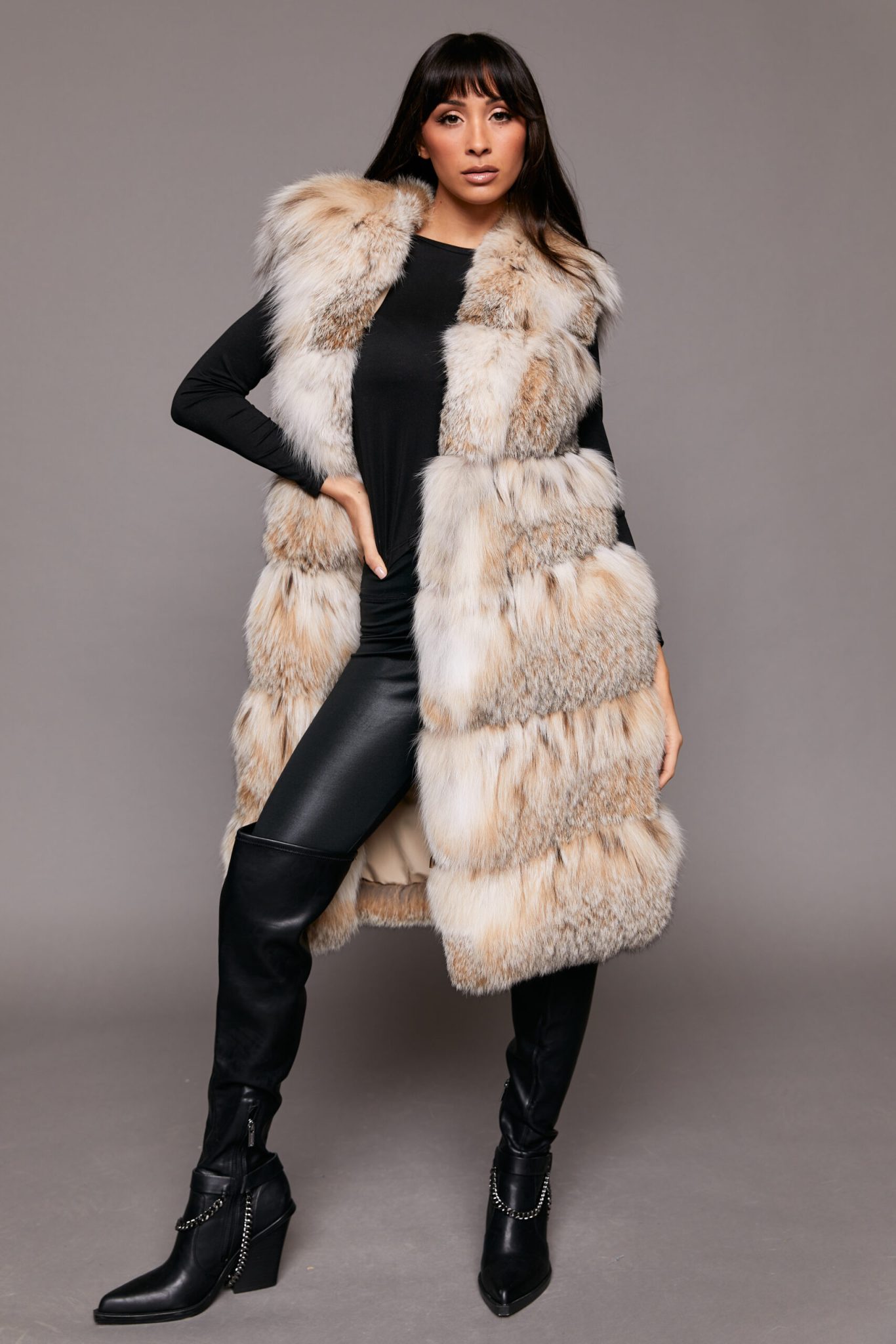 Full length shot of a woman wearing a fur vest standing with her right hand on her hip in front of a grey background