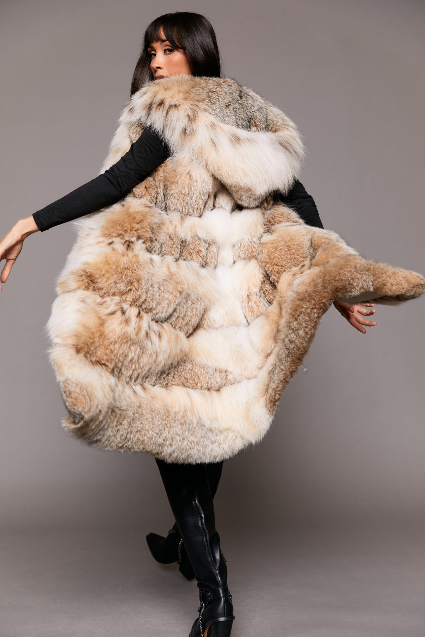 Full length shot of a woman wearing a fur vest twirling so the lower part of the vest is fluttering in the air, standing in front of a grey backdrop